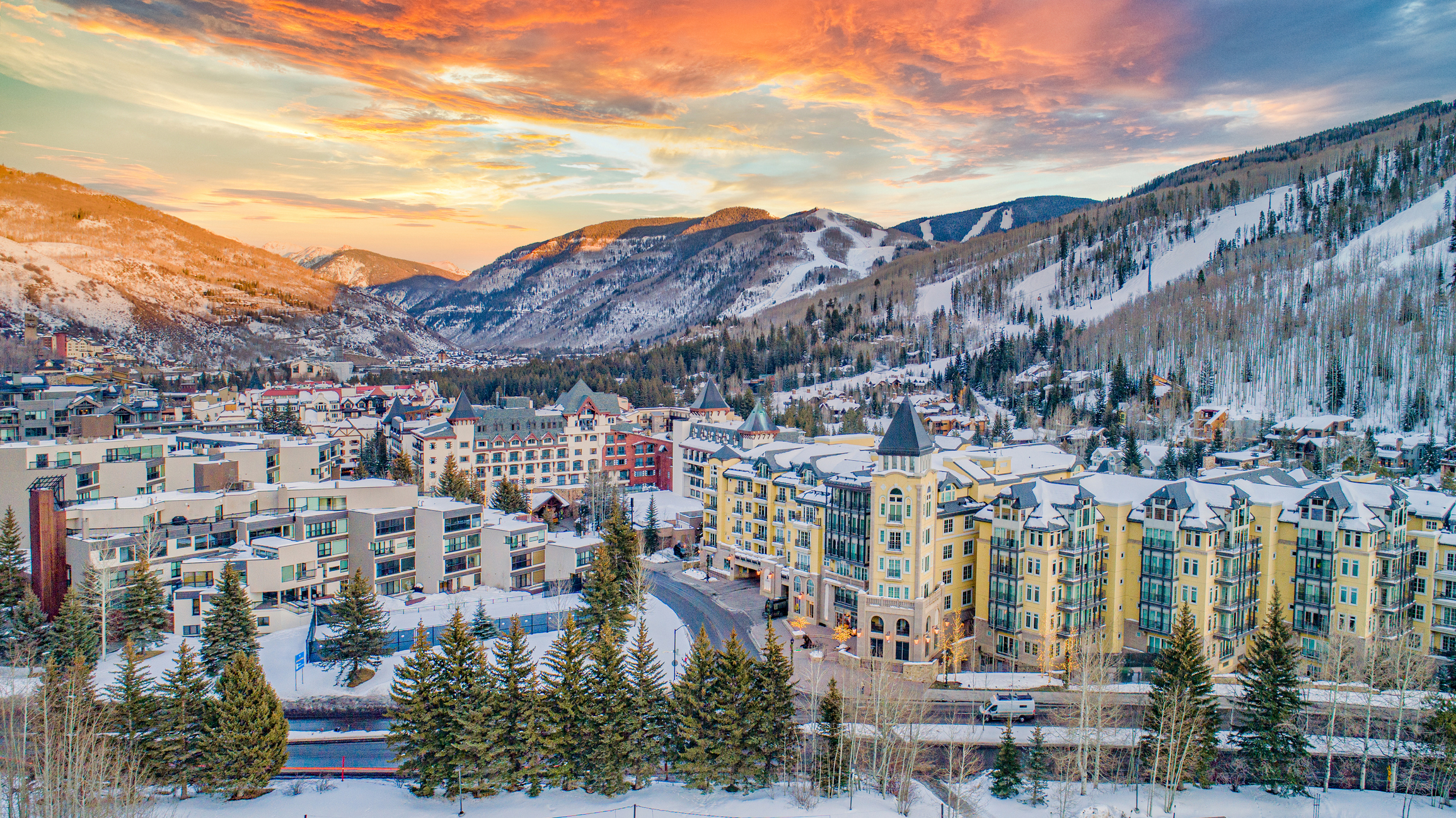 Scenic winter view of a town nestled in snow-covered mountains with colorful buildings and a dramatic sunset sky in the background