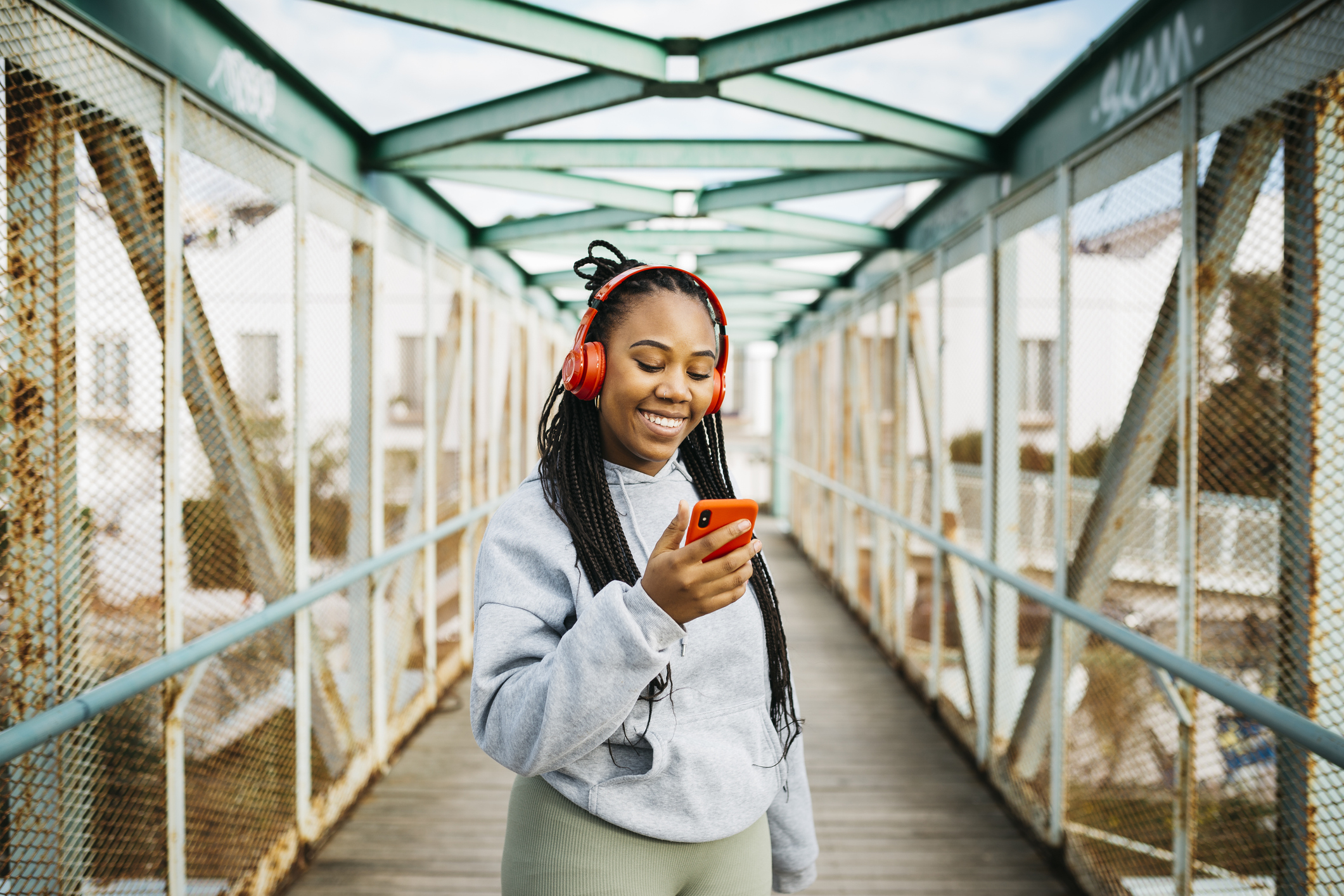 A woman smiles while looking at her phone and listening to music on a pedestrian bridge.