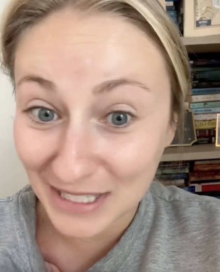 A woman with a headband and grey top smiles and speaks in front of a bookshelf