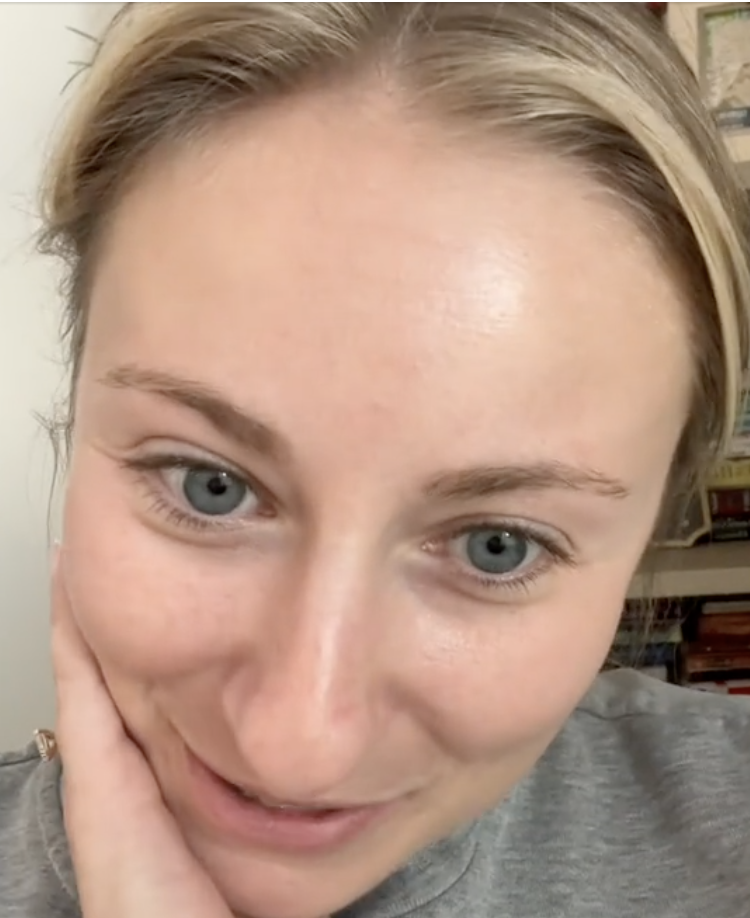 Close-up of a woman with her hand on her cheek, smiling at the camera. She is wearing a simple top