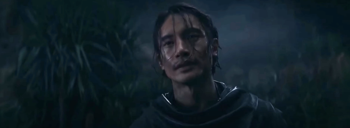 A man with wet hair and a solemn expression stands in a dark, misty forest, wearing a cloak