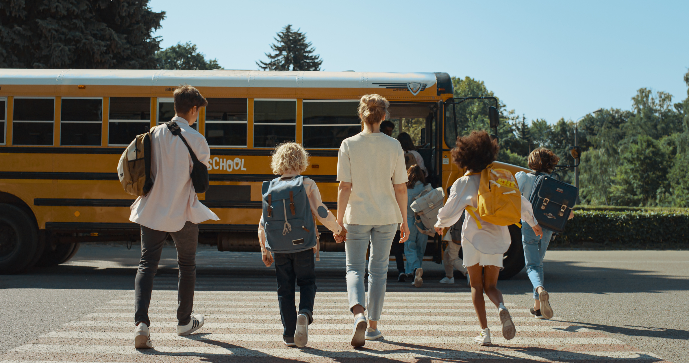 Five children with backpacks walk towards a school bus. They appear joyful, heading towards an adventure or school day. Trees are visible in the background
