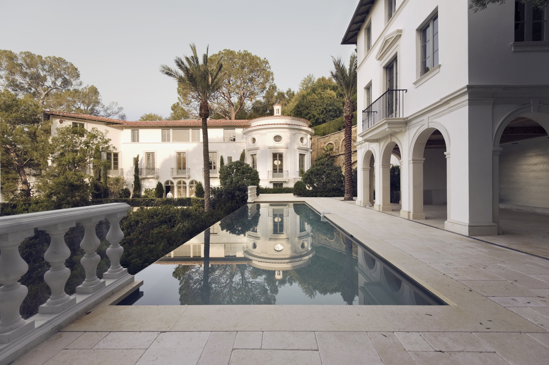 Large luxury mansion with arched walkways and a long reflecting pool in the front yard, surrounded by trees