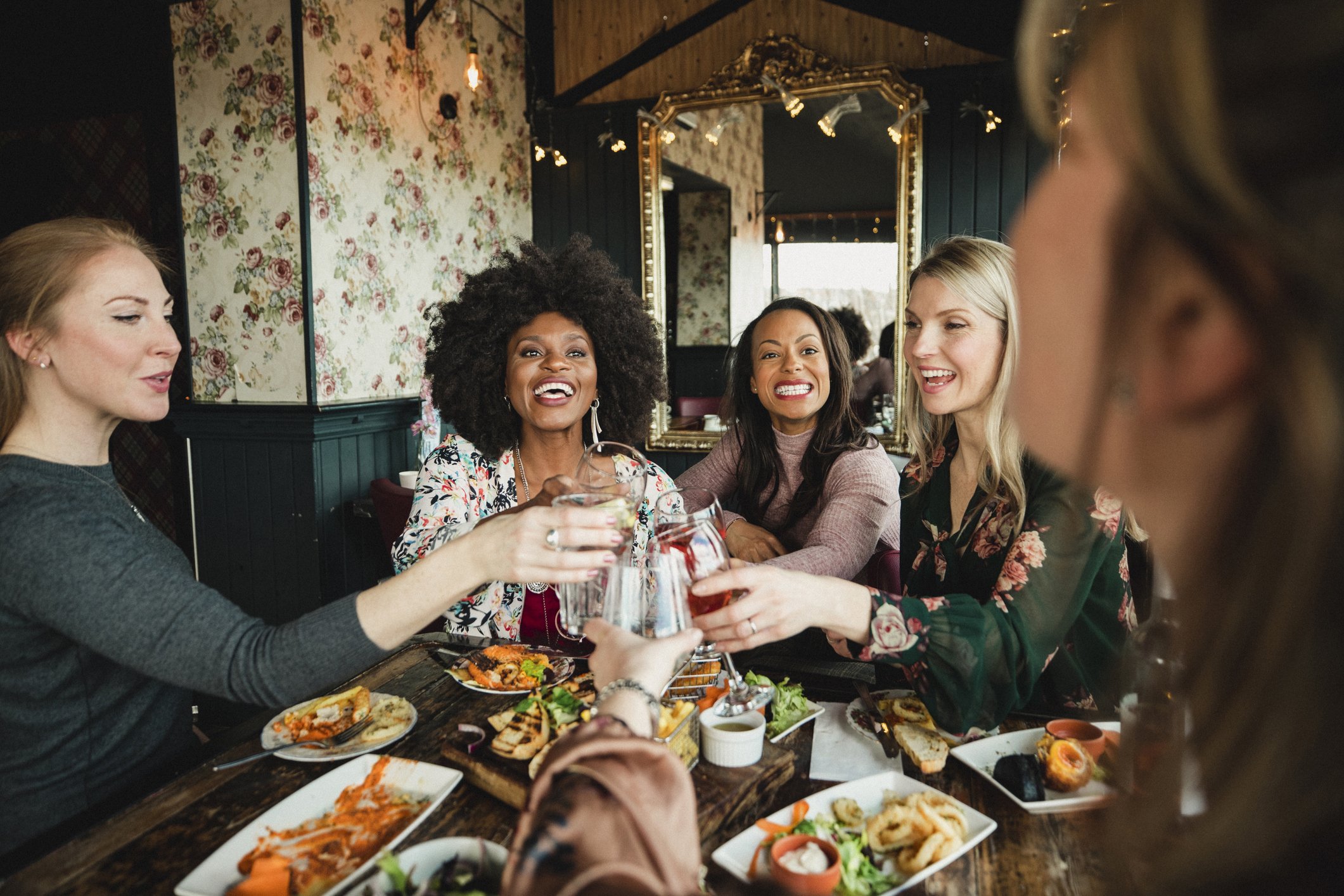 A group of five women at a cozy restaurant, smiling and toasting with drinks over a table full of food, enjoying a cheerful moment together