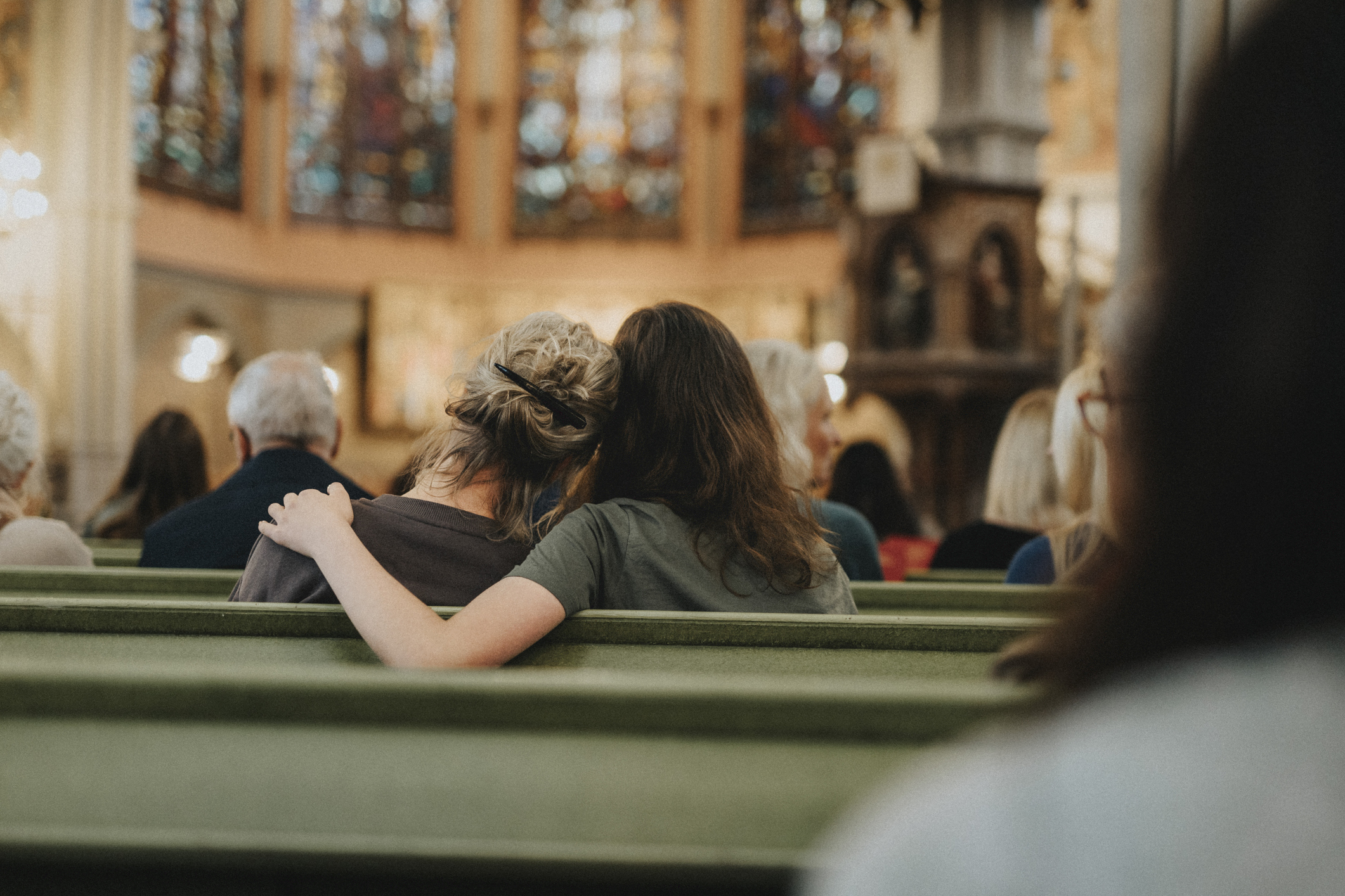 Two people sit closely in a church pew; one has an arm wrapped around the other in a comforting gesture. Faces are not visible