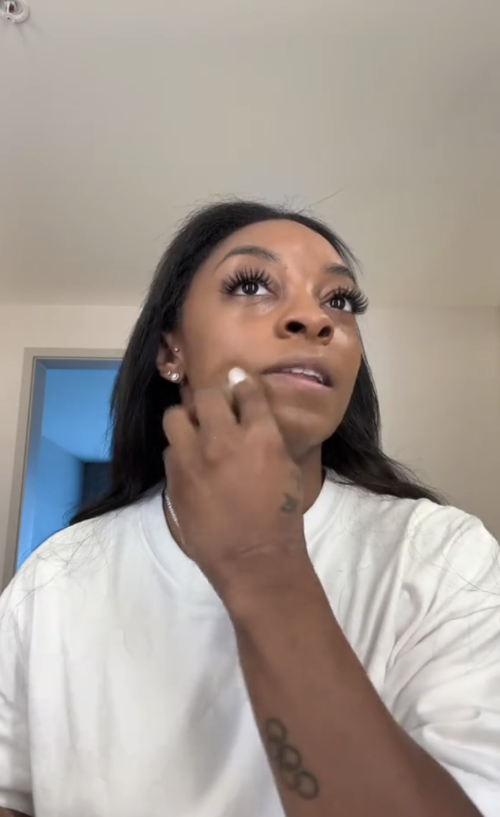 Simone is applying makeup to her face, wearing a shirt. She stands in a neutral room, with a slightly open door visible in the background