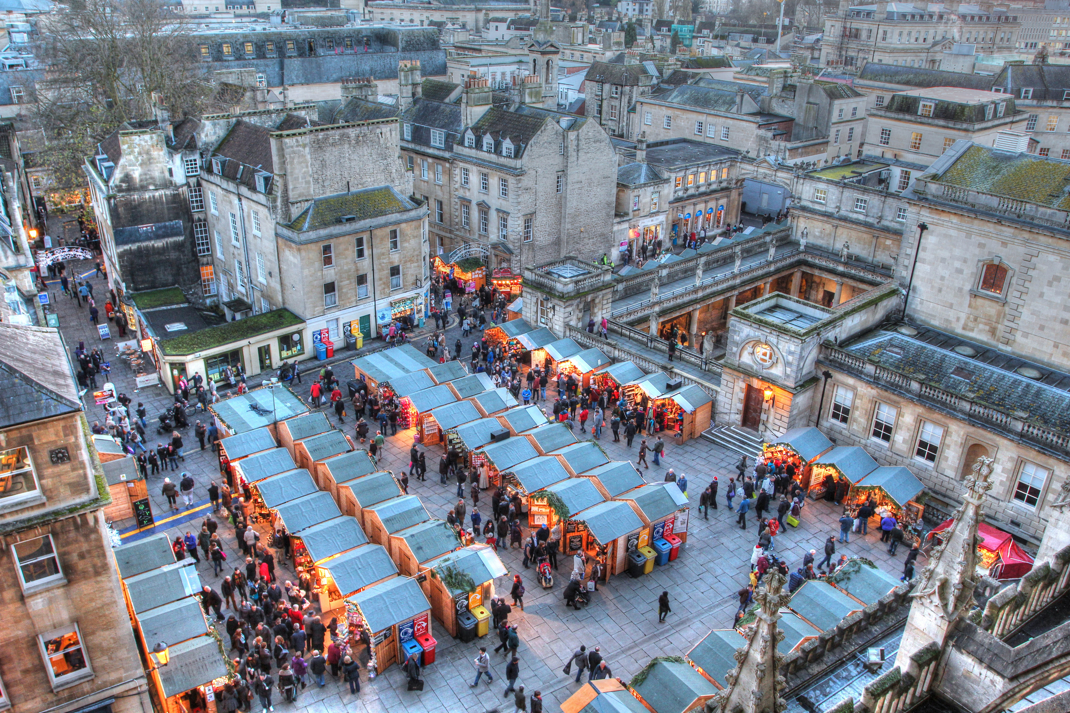 A bustling outdoor market with numerous stalls and many people browsing. The market is set in an urban area surrounded by historic buildings