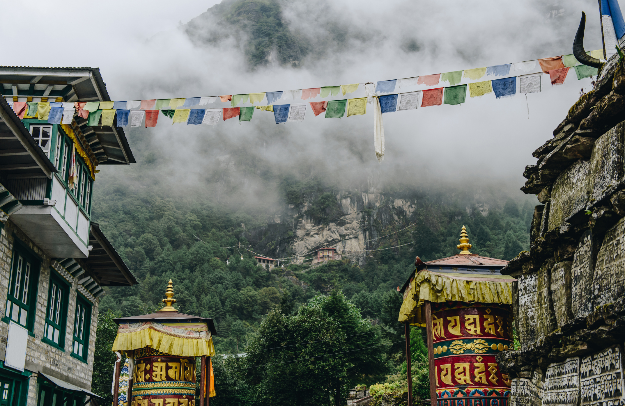 Mountain village scene with traditional prayer flags, buildings with intricate details, and stone structures amid misty, forested mountains. No people present