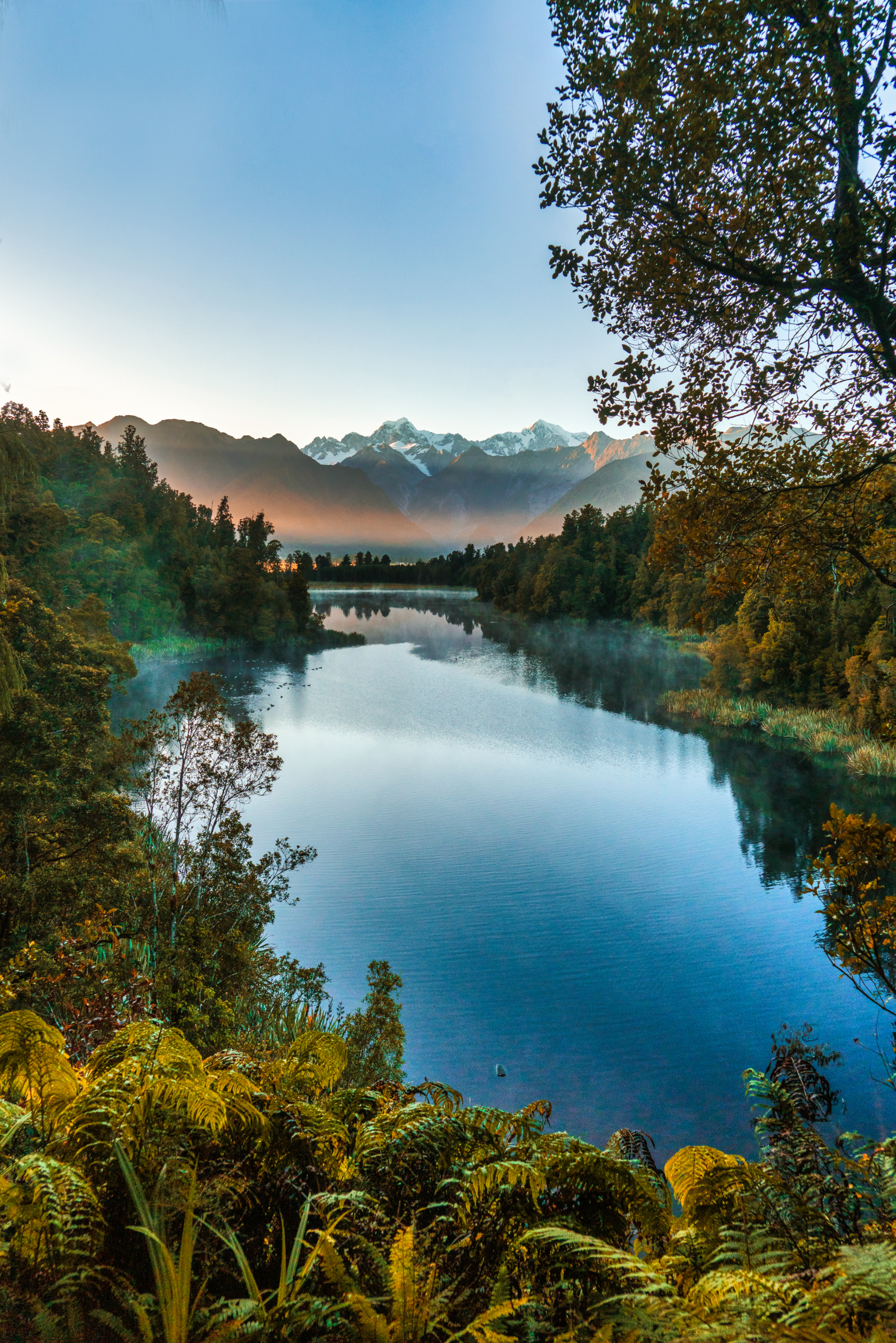 A serene lake surrounded by lush trees with snow-capped mountains in the background under a clear sky