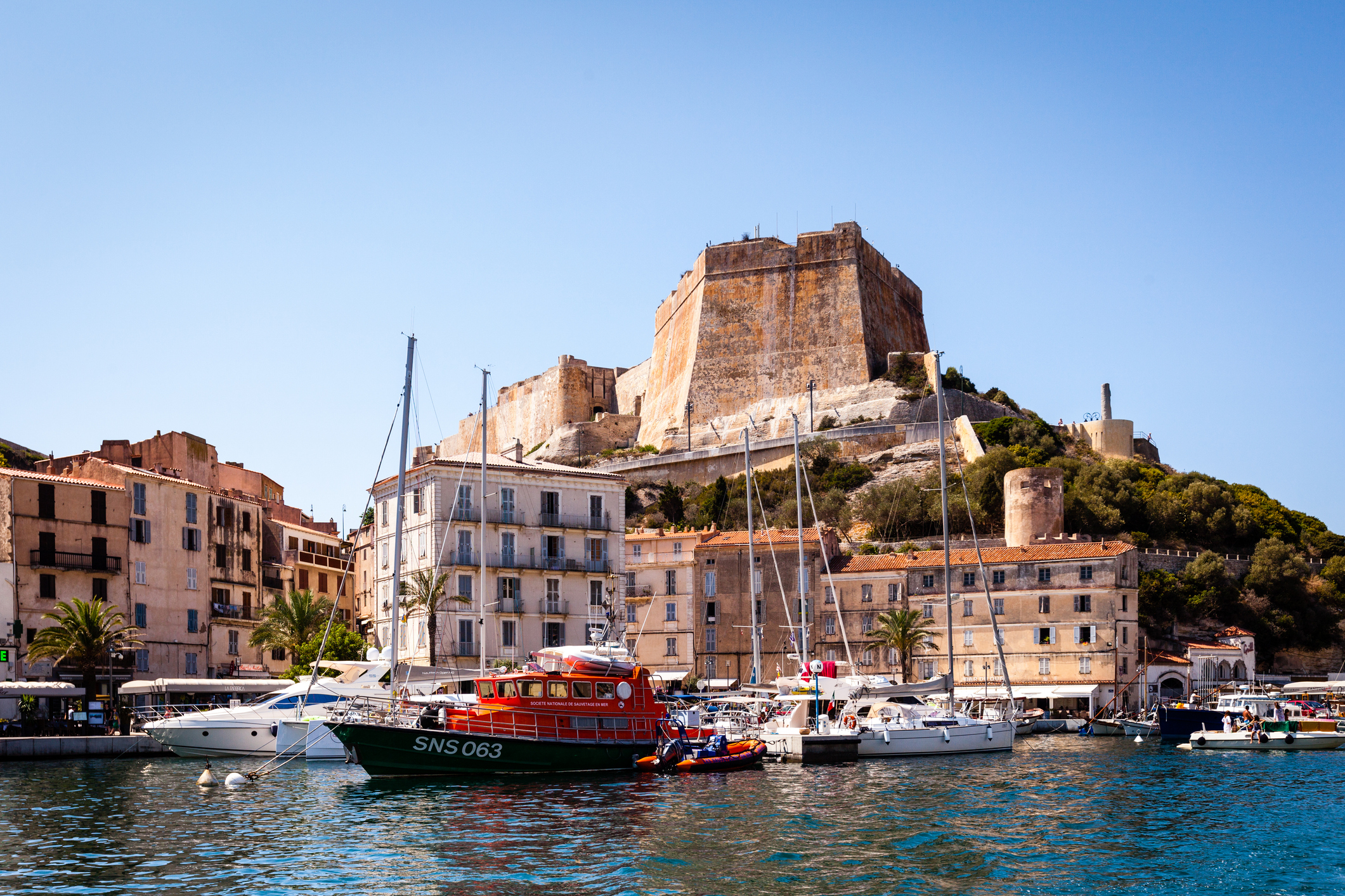 Boats docked in a harbor with a historic stone fortress on a hill in the background, and multi-story buildings along the waterfront