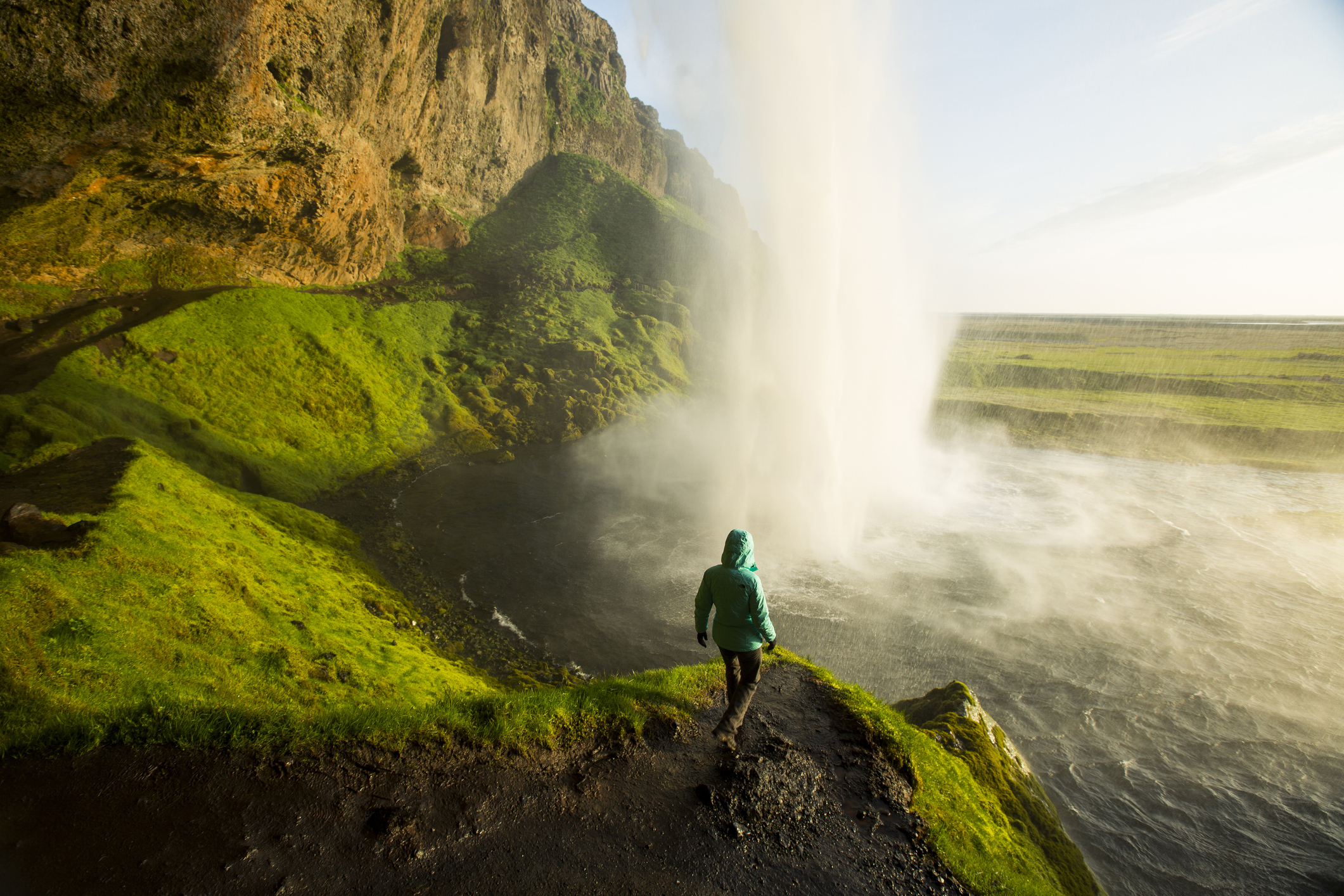 Person in a hooded jacket standing on a trail near a large waterfall, with vibrant greenery and cliffs in the background scenery