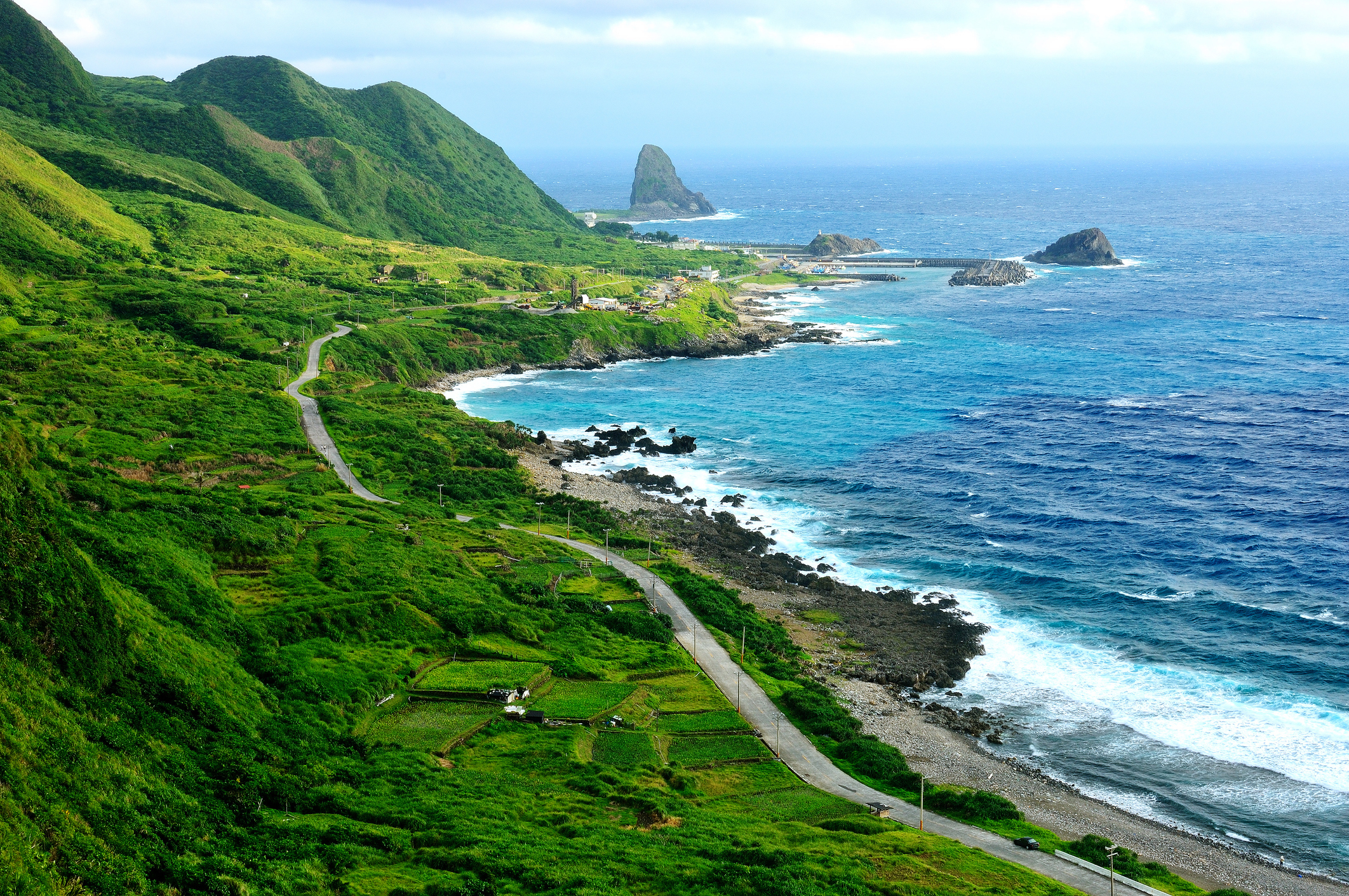 Coastal landscape with winding road, lush green hills, rocky shoreline, and ocean waves. There&#x27;s no text or people in the image