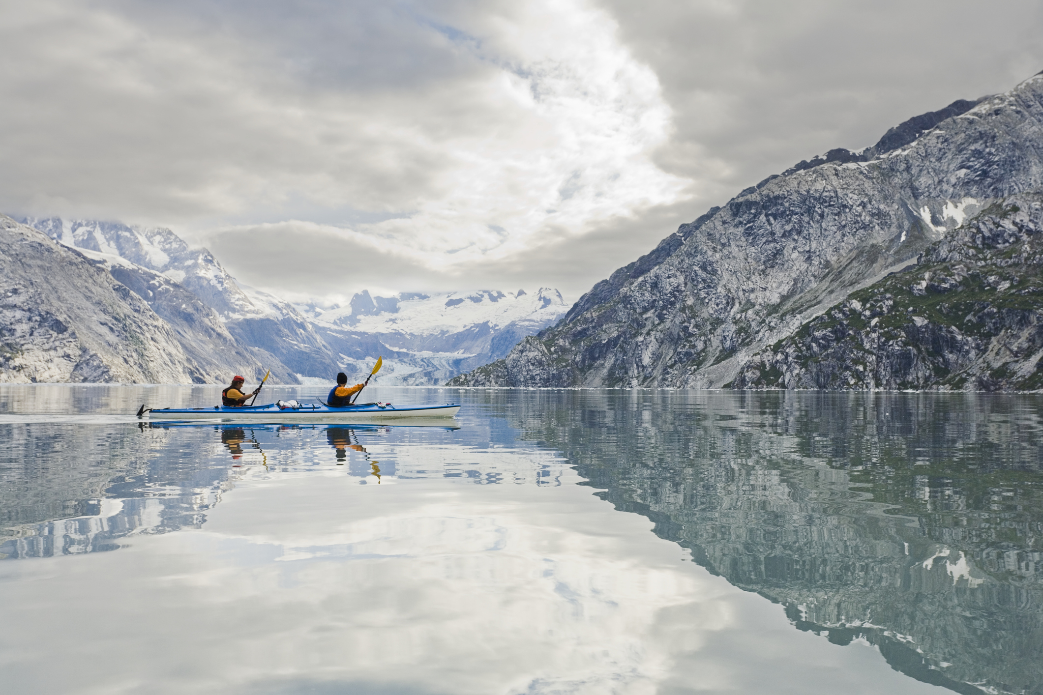 Two people kayaking on a calm mountain lake surrounded by snow-capped peaks and lush slopes under a cloudy sky