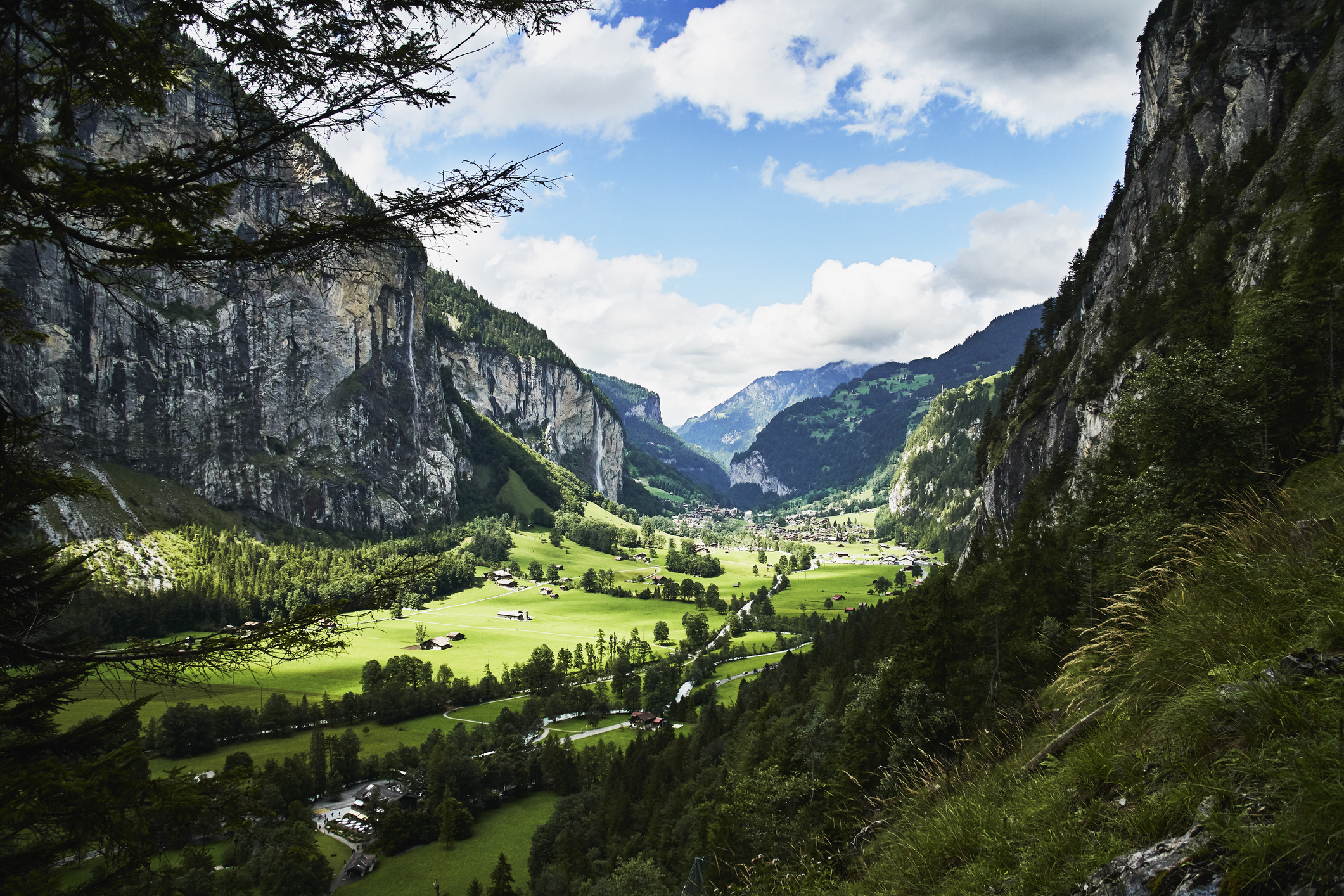 A scenic view of a lush green valley surrounded by mountains, with scattered houses and a small village in the distance under a partly cloudy sky