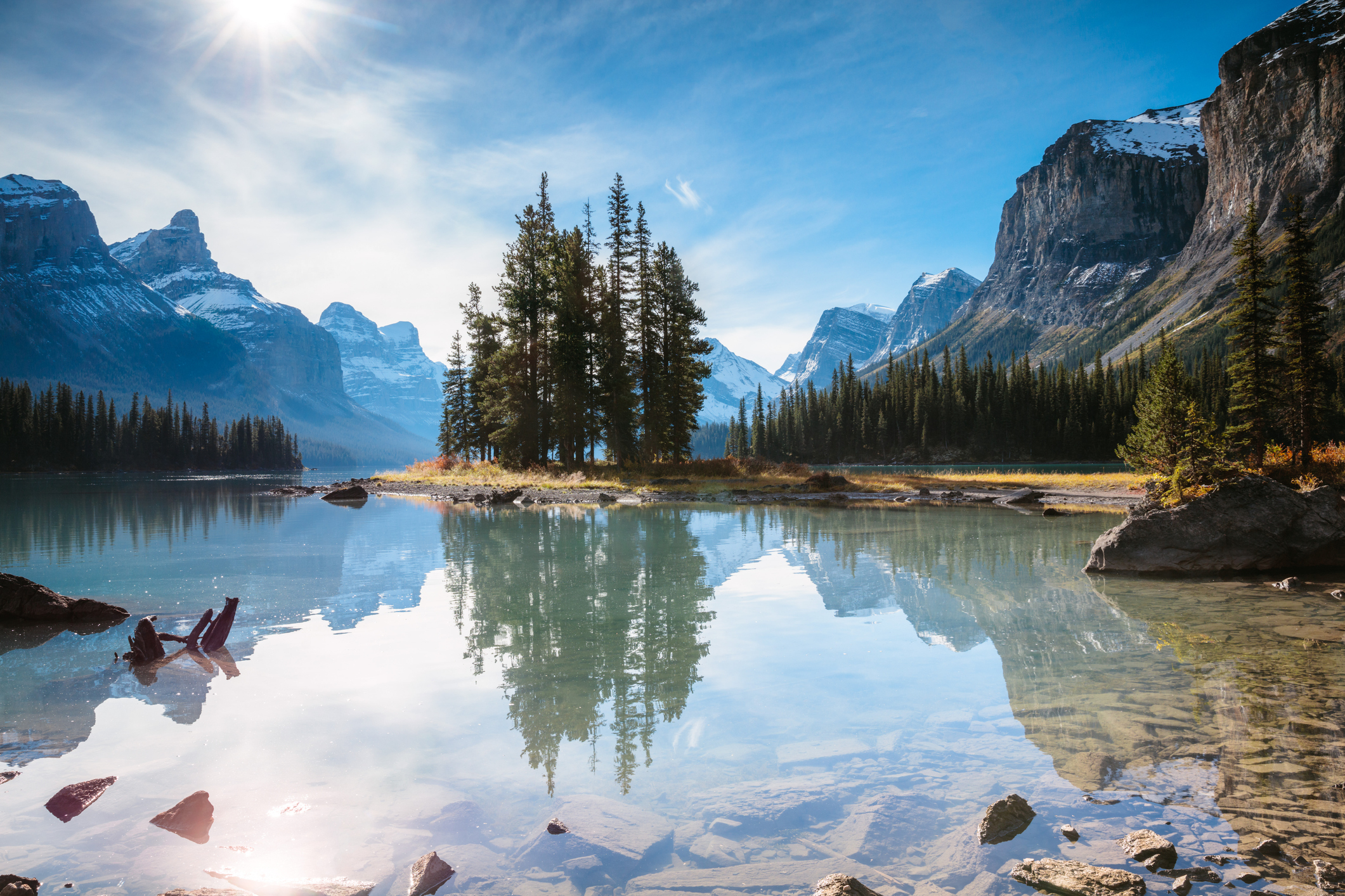 A serene lake surrounded by mountains with a cluster of trees on an island in the middle. The sky is clear, and the sunlight illuminates the scene