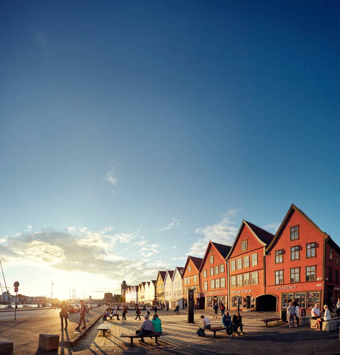 People strolling and sitting along a waterfront with a row of distinctive, steep-roofed buildings in the background under a clear sky