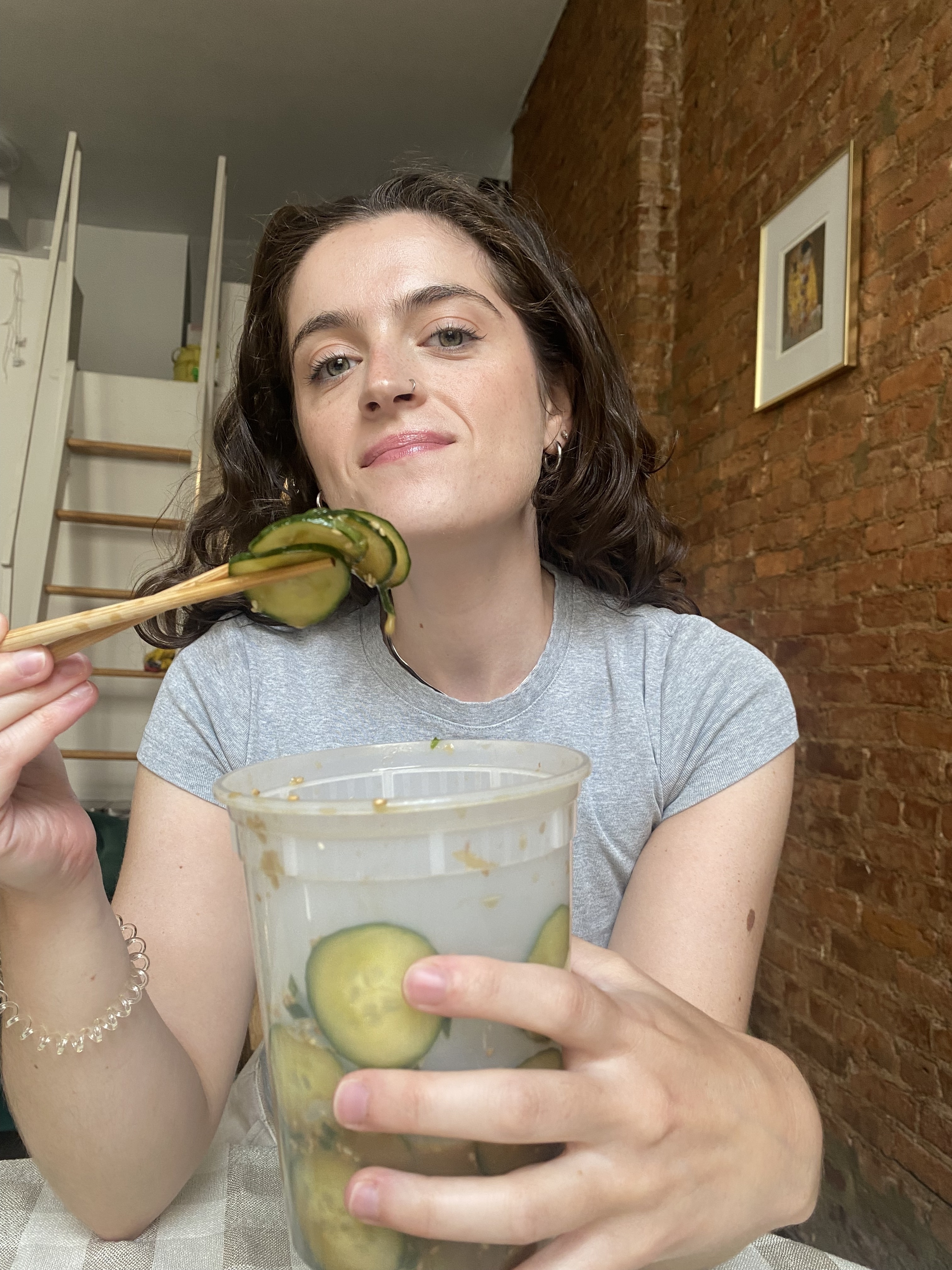 A woman in a grey top is using chopsticks to eat pickled cucumbers from a large container in a kitchen