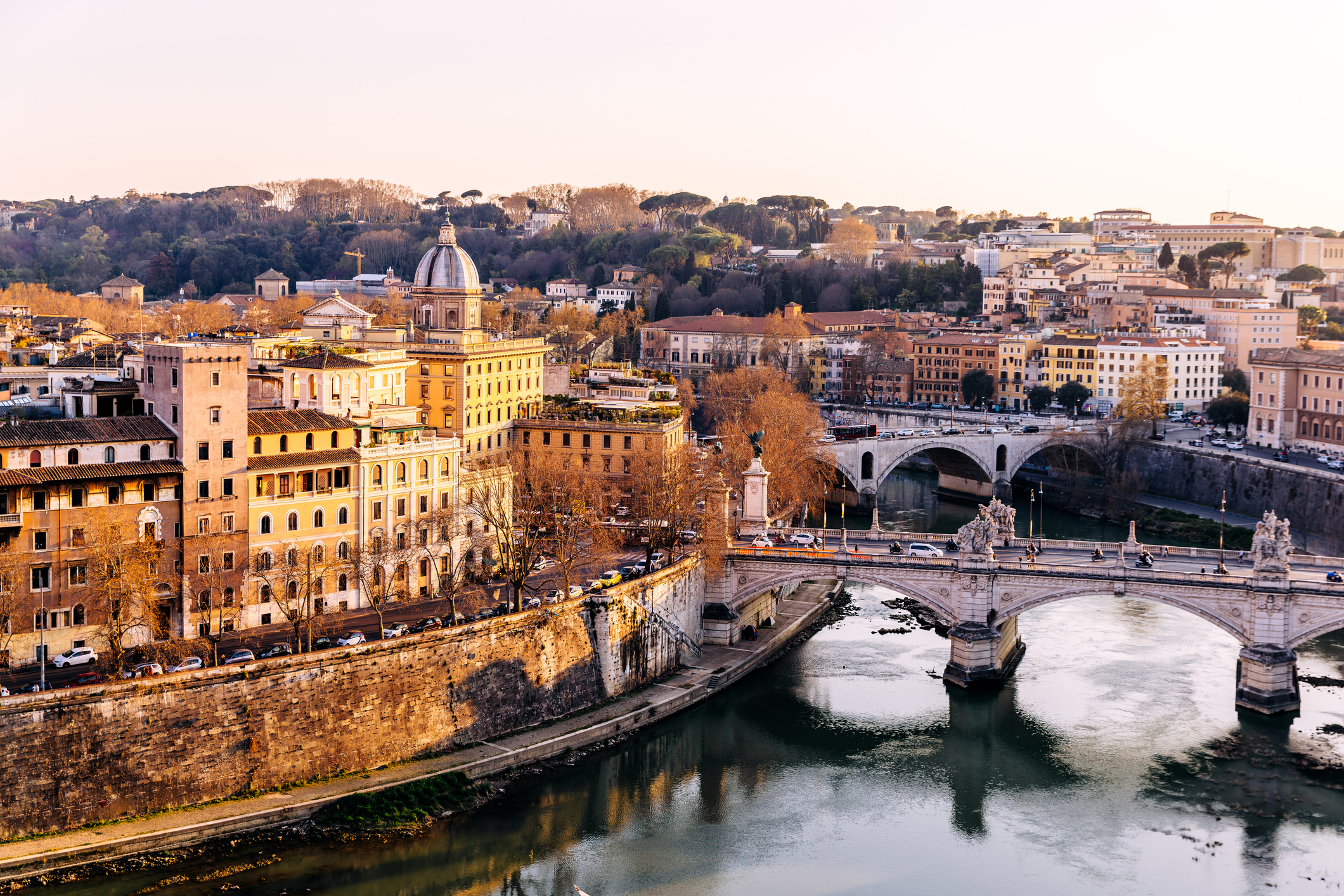 A scenic view of Rome, Italy, featuring historic buildings, the Tiber River, and the Ponte Sant&#x27;Angelo bridge. Trees and hills frame the background