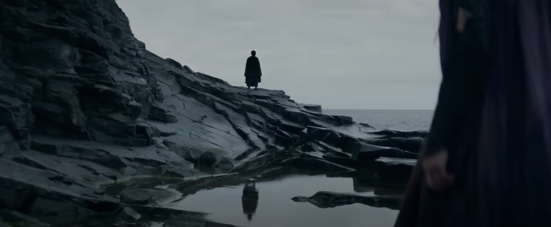 A lone figure stands on rocky cliffs overlooking the sea,