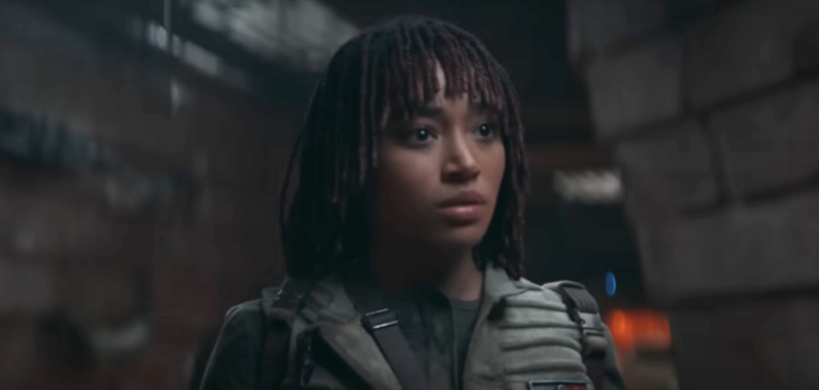 Amandla Stenberg in rugged, sci-fi clothing with a stern expression, standing in a dimly lit, industrial setting