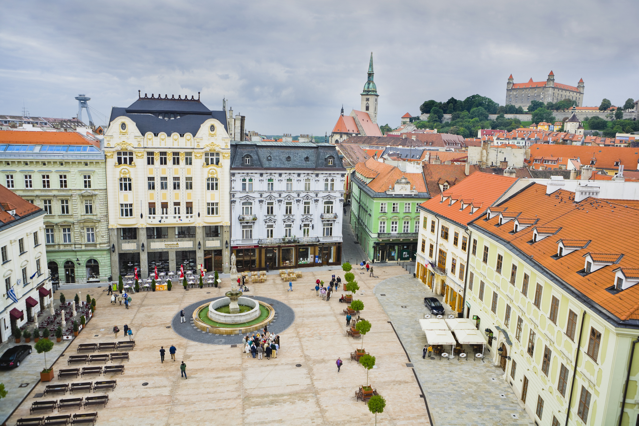 Aerial view of a European town square with historic buildings surrounding a central fountain and scattered pedestrians. Treed hills and castle in the background