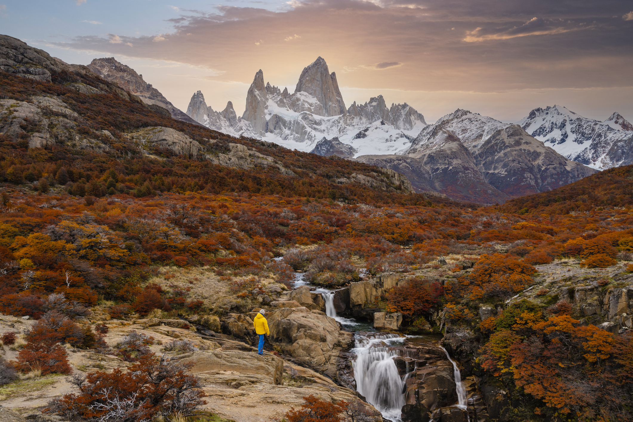 A lone hiker stands on a rocky terrain wearing a yellow jacket, overlooking a waterfall with towering mountains and a dramatic sky in the background