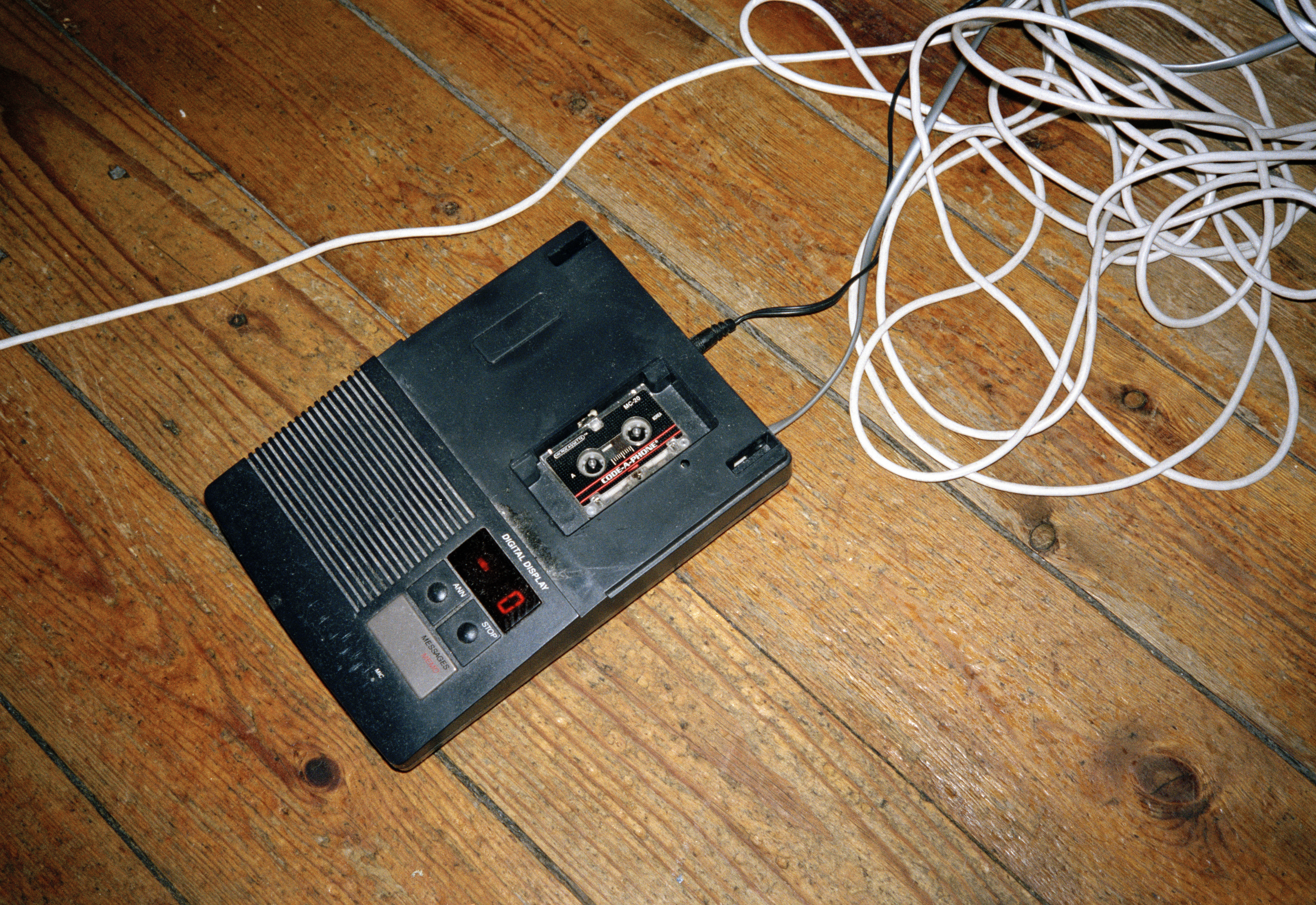 Vintage cassette tape recorder on a wooden floor, connected to a tangled white cord