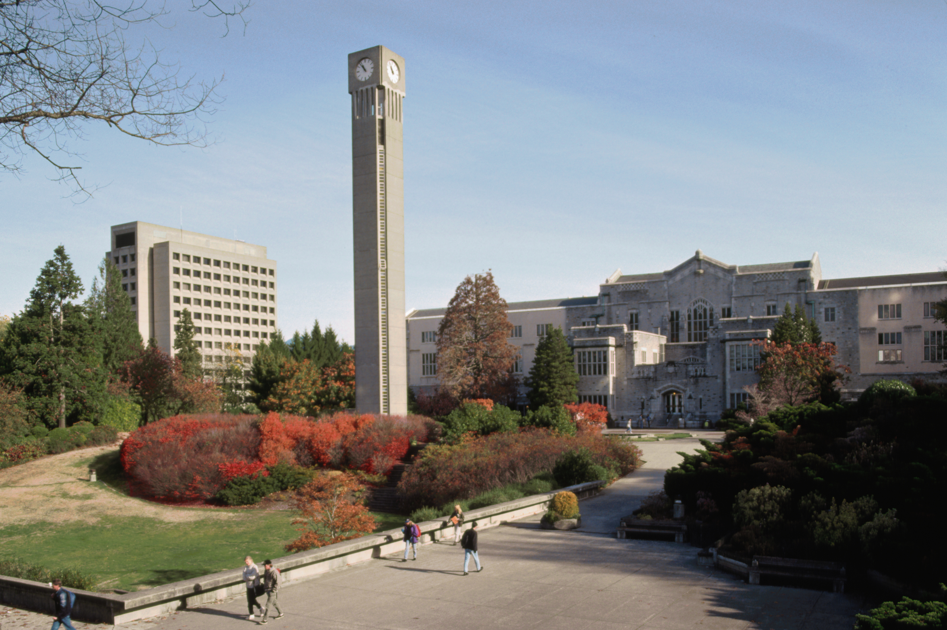 A university campus featuring a tall clock tower, modern and gothic-style buildings, and students walking on pathways surrounded by landscaped greenery