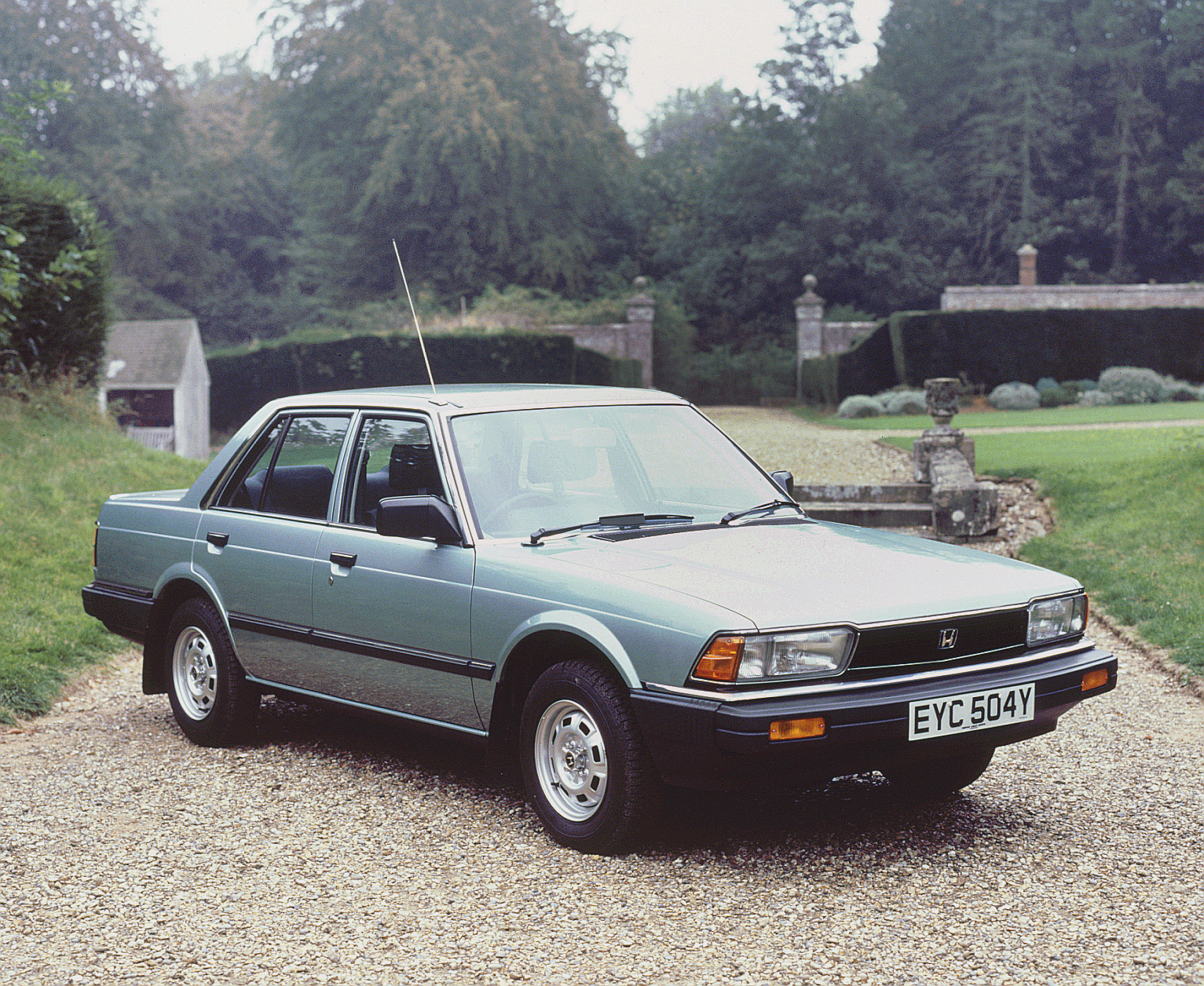 A classic Honda Accord sedan parked on a gravel driveway in a countryside setting, with trees and a brick structure in the background. License plate reads EYC 504Y