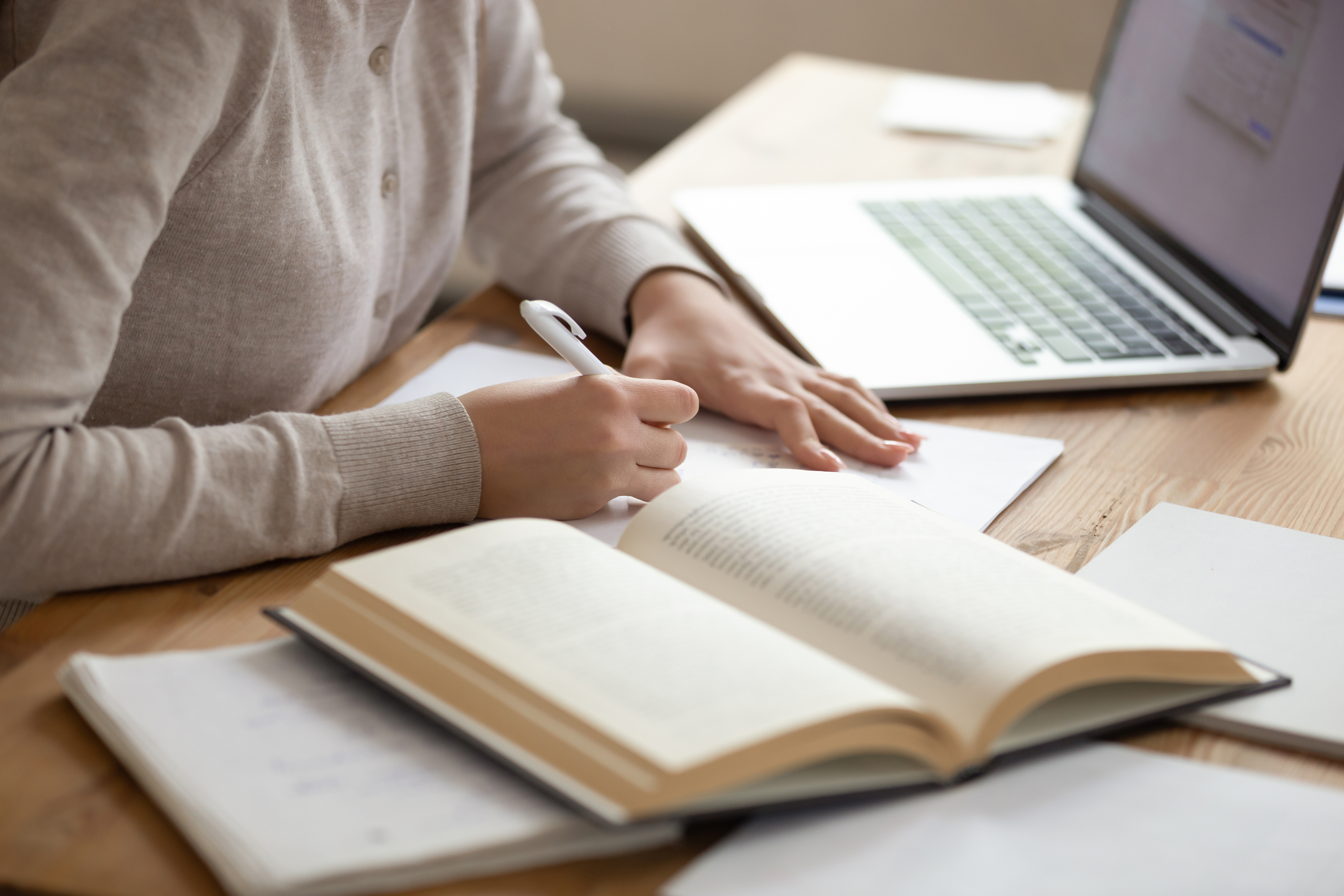 A person writes in a notebook with an open book and a laptop on a desk, suggesting a study or work session