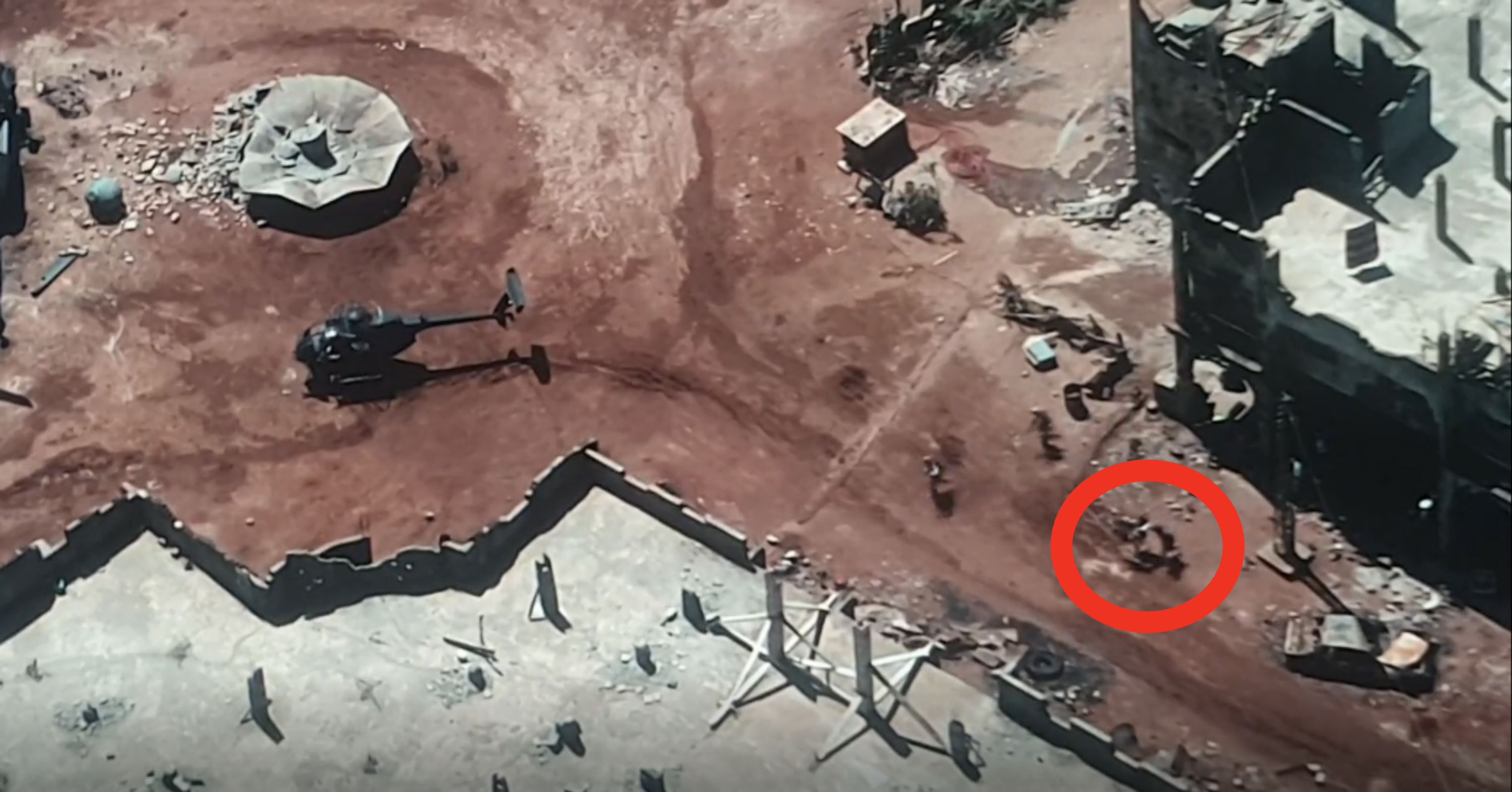 A helicopter lands on a dirt ground in the midst of a rugged, partially destroyed urban area with debris and buildings visible. Multiple people can be seen nearby