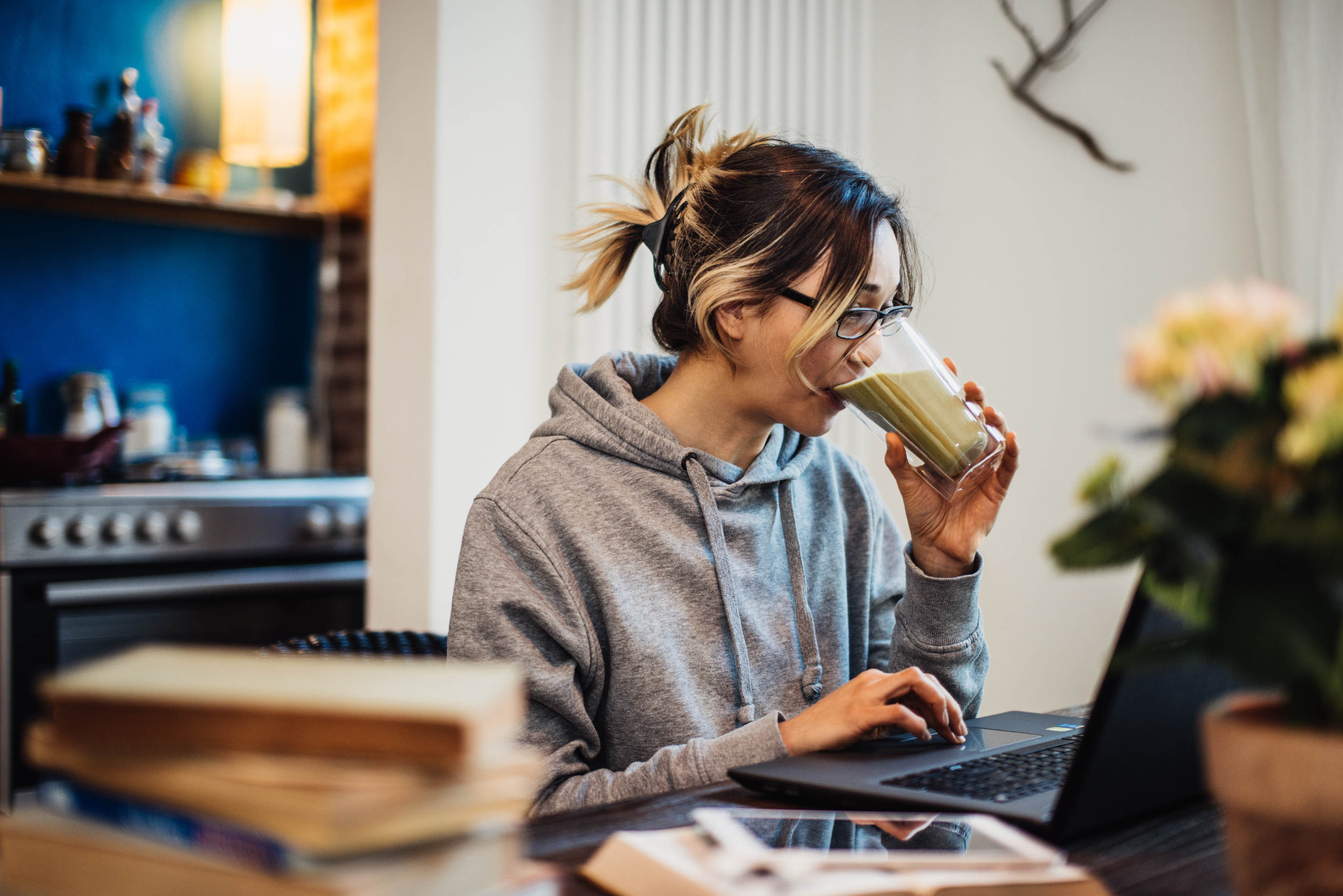 A person is sitting in front of a laptop in a cozy kitchen, drinking a smoothie and wearing glasses and a hoodie. Books and a smartphone are on the table