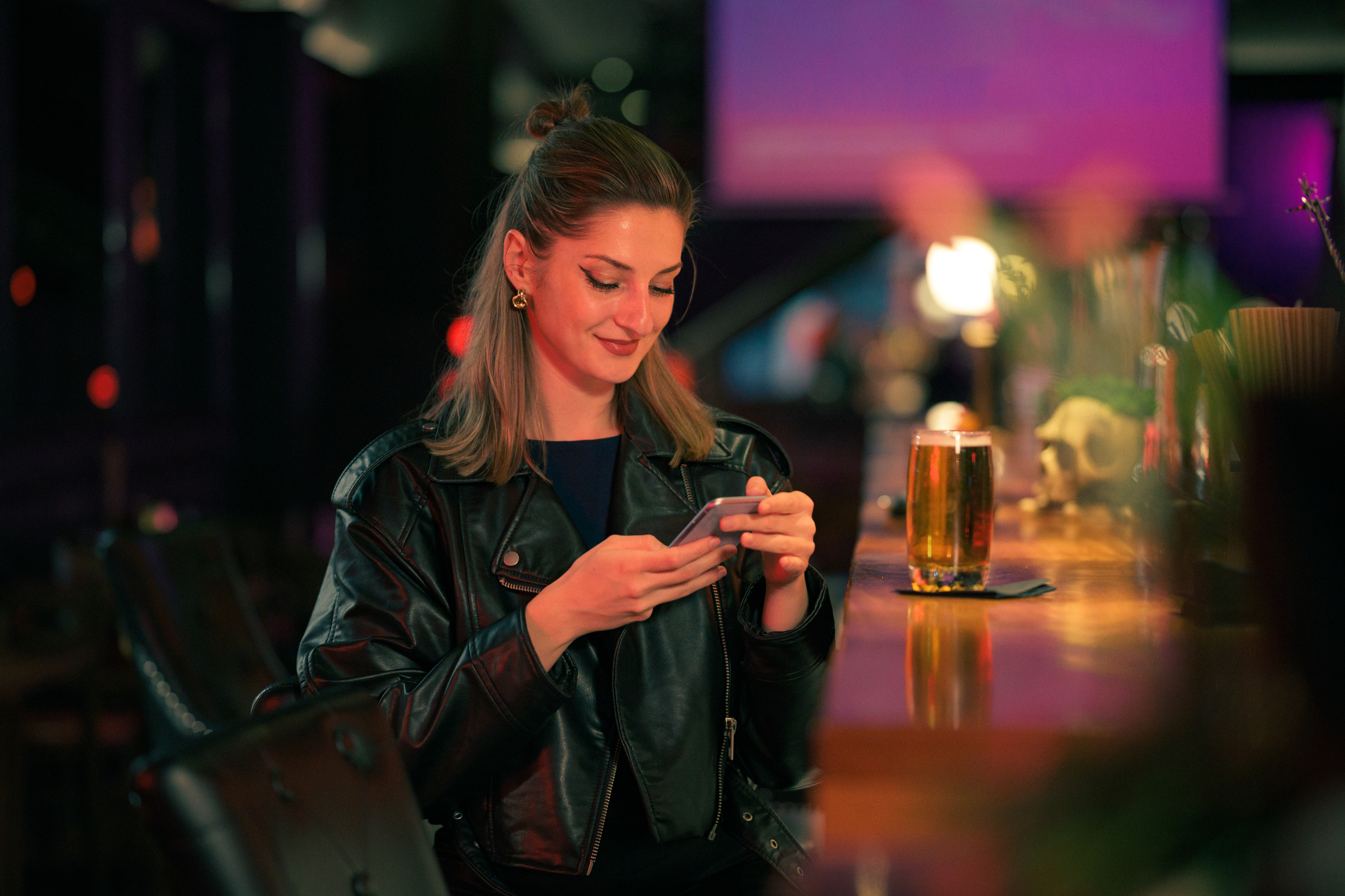 A woman in a stylish leather jacket sits at a bar, looking at her phone. A glass of beer is in front of her