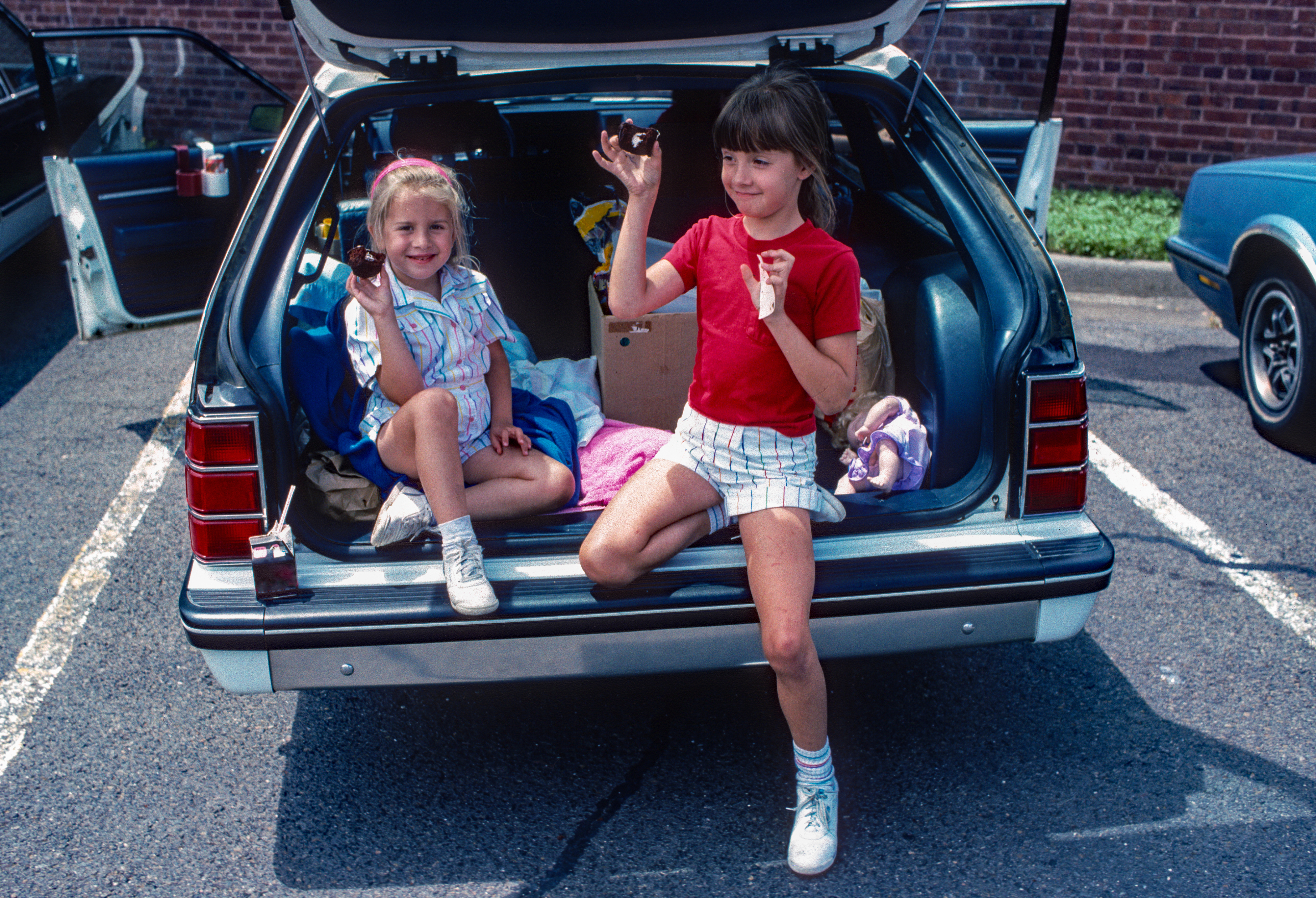 Two young girls, presumably siblings, sit in the trunk of a station wagon. One wears a blue and white dress, the other a red shirt and striped shorts