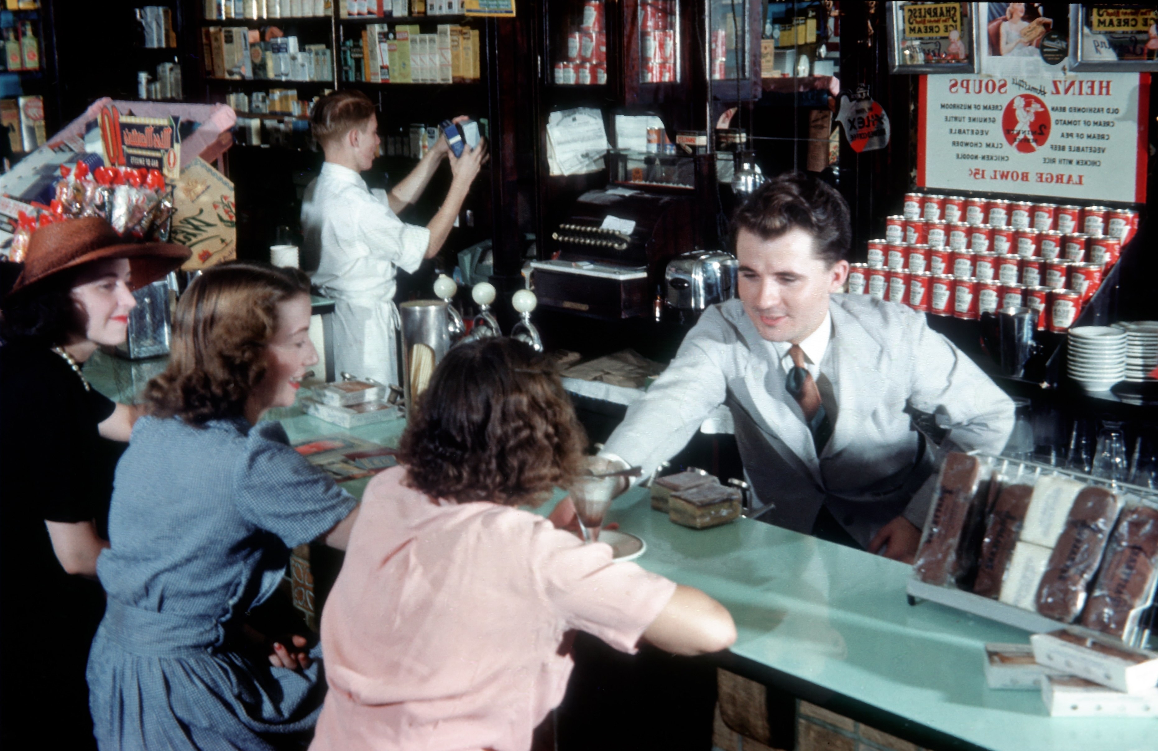 A 1940s soda fountain scene with a man in a white coat serving ice cream to three patrons at the counter
