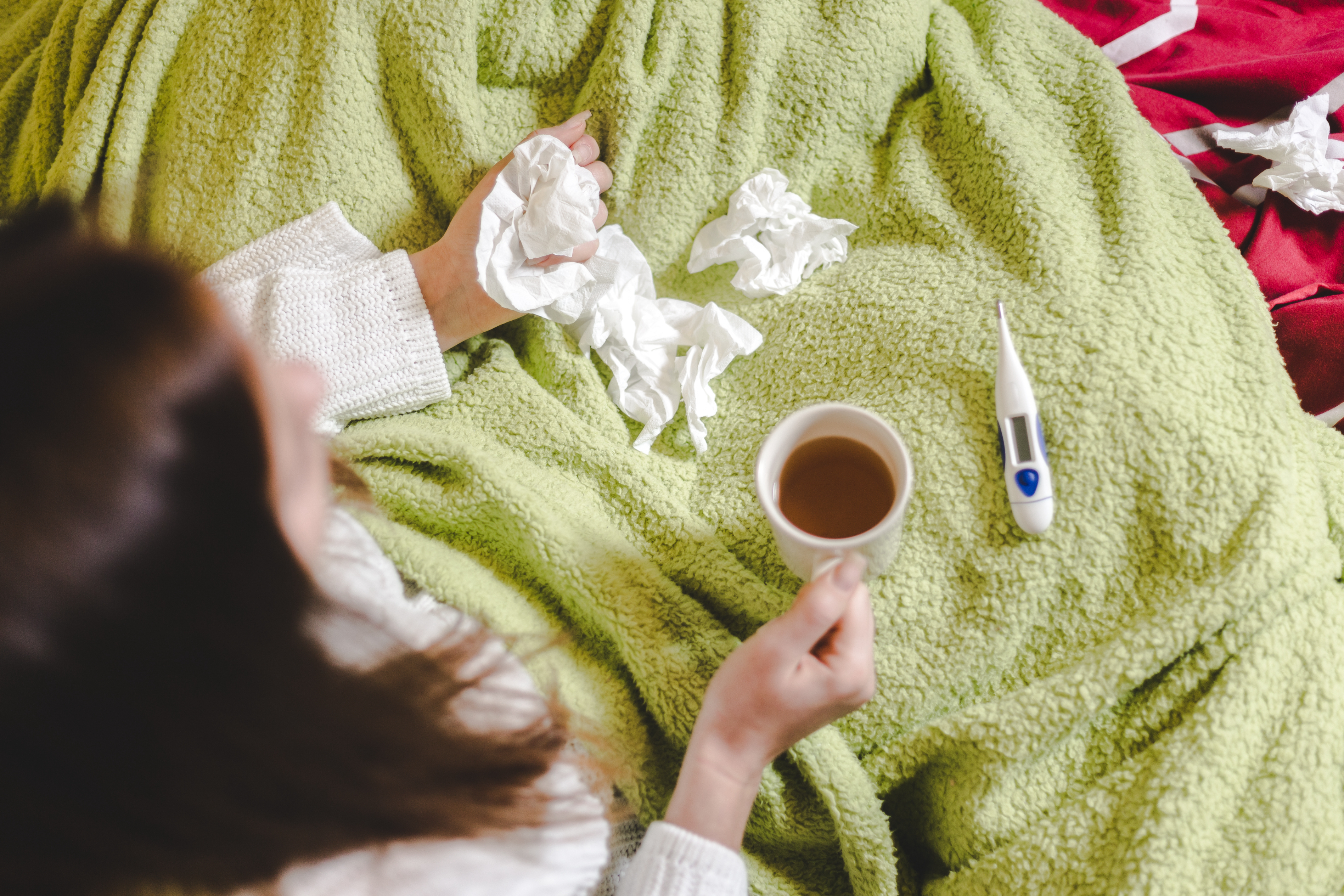 A person holding a coffee cup and a tissue, sitting under a blanket with a thermometer nearby, possibly feeling unwell