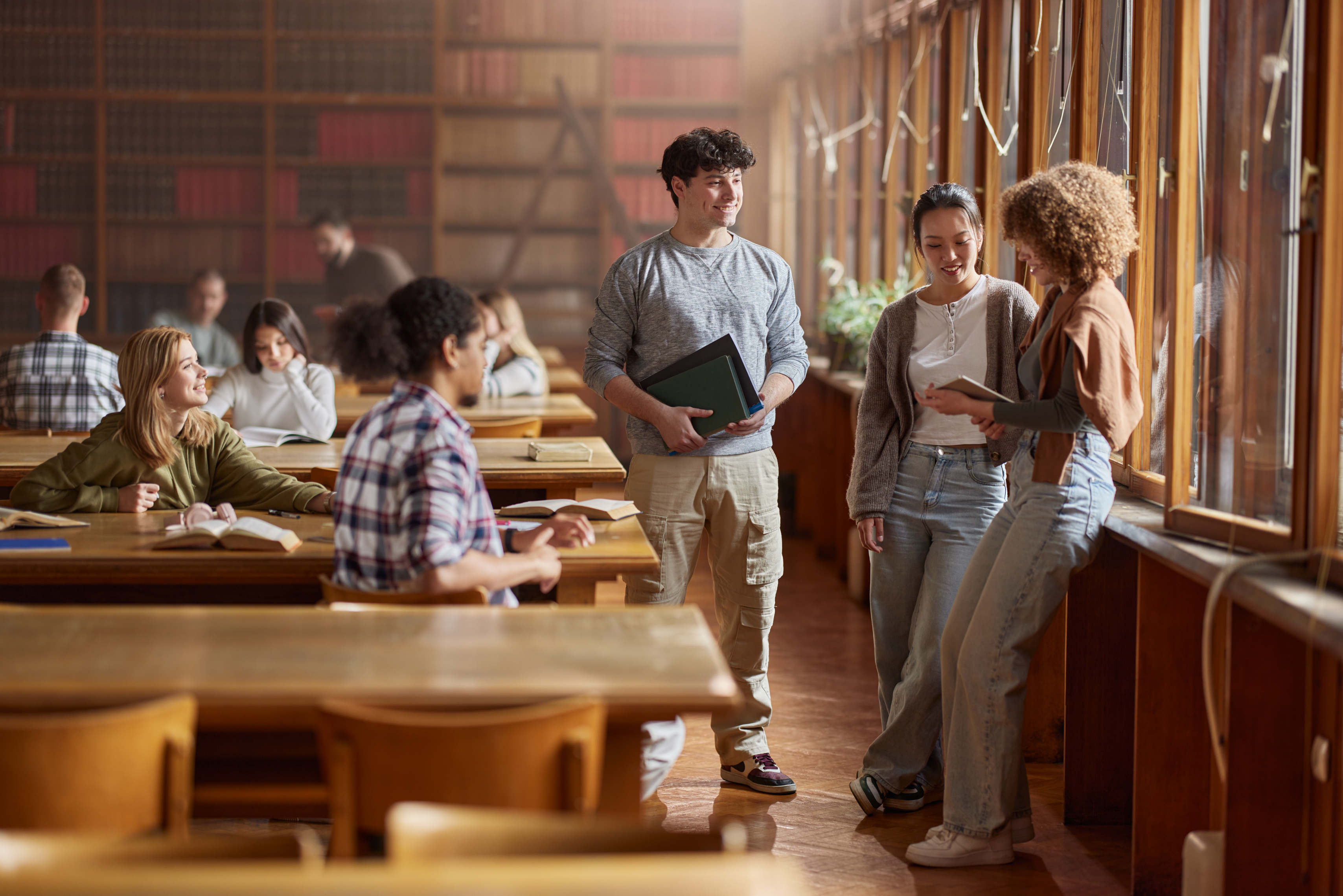 A group of students, including a person holding a folder, stands and sits in a library, engaged in conversation and studying. The room is filled with shelves of books