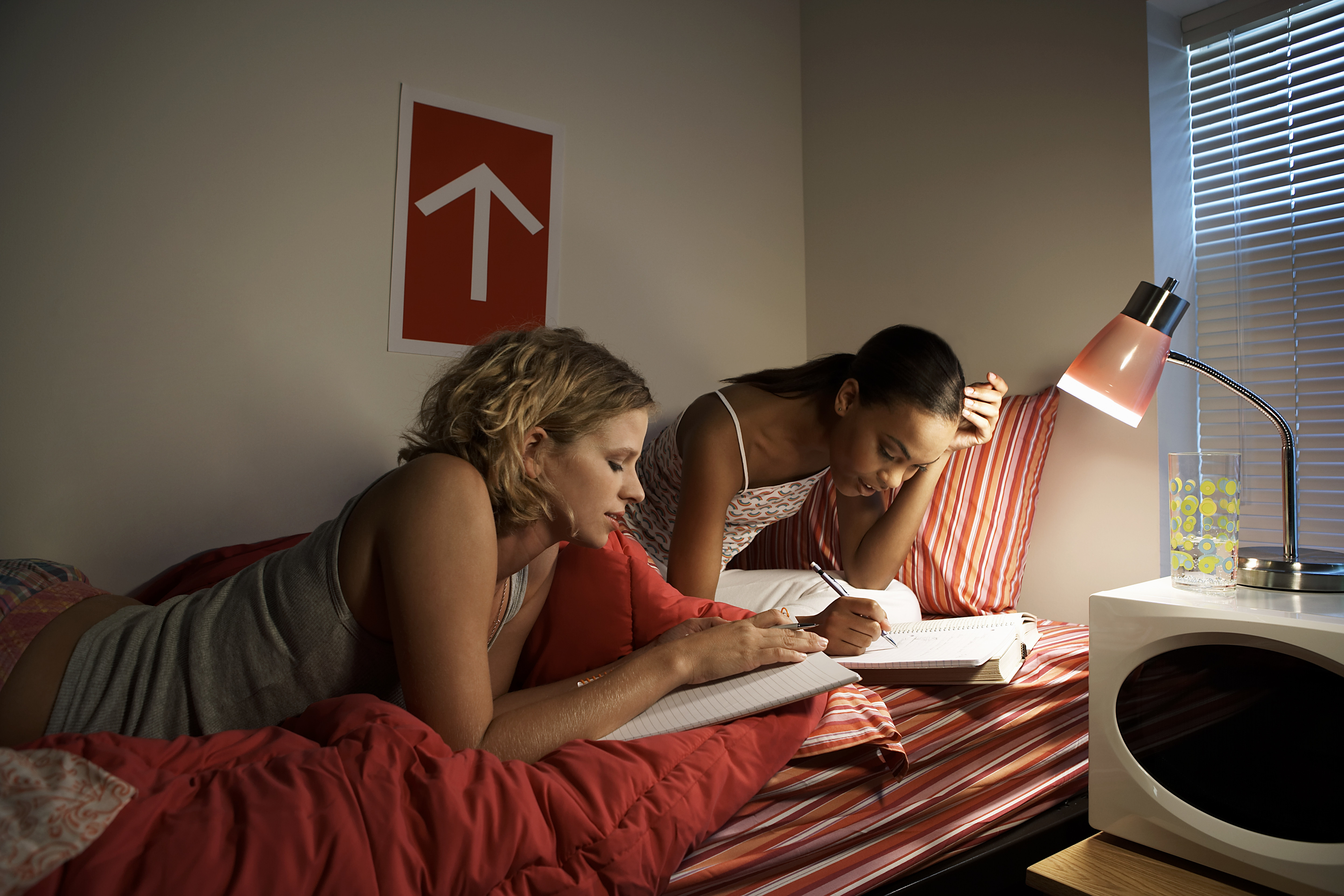 Two women study together on a bed under a lamp. One writes in a notebook, the other looks on. A red sign with an upward arrow is on the wall behind them