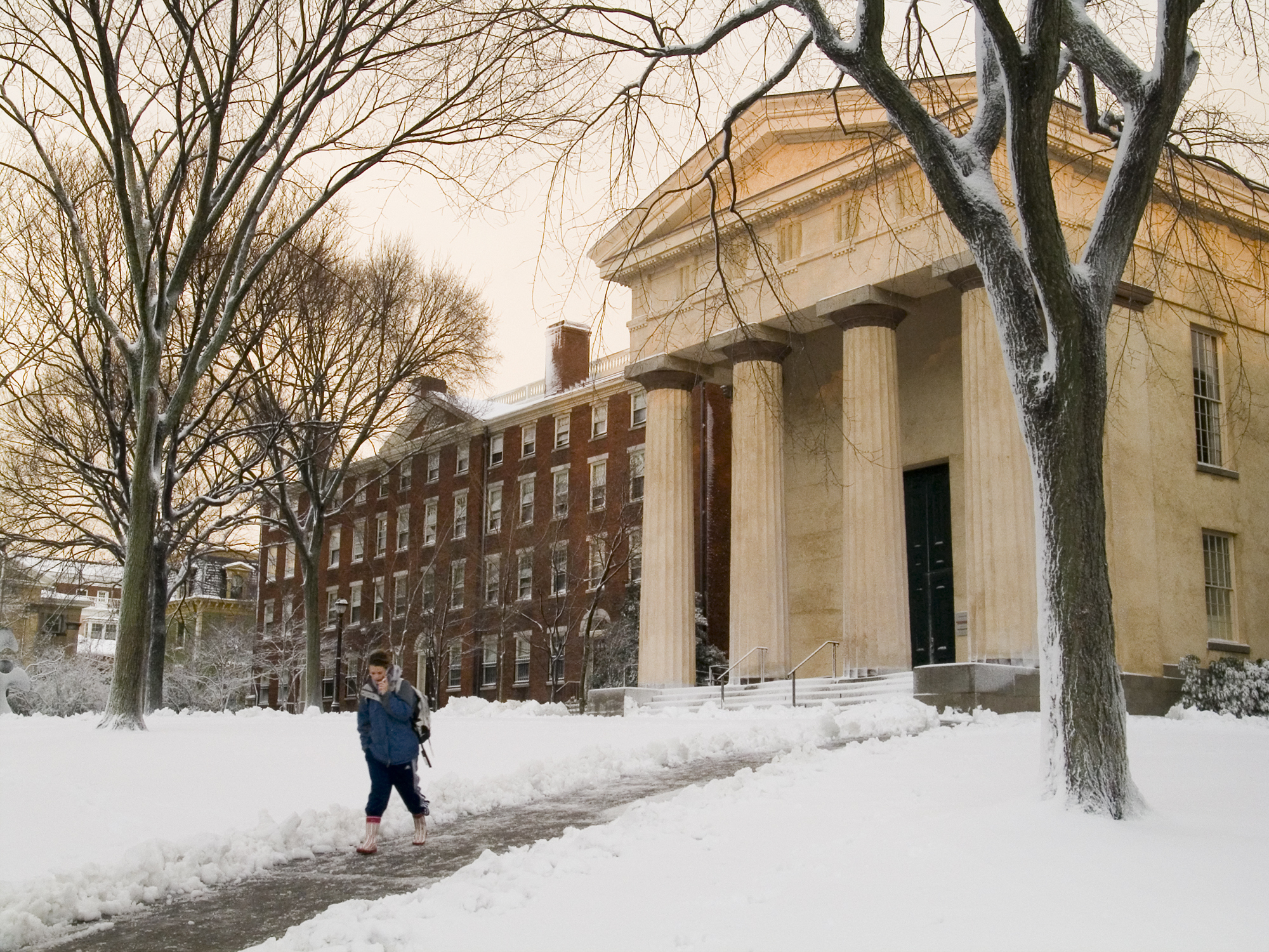 Person walks down a snowy path past a historic university building with columns and a row of red brick buildings in the background