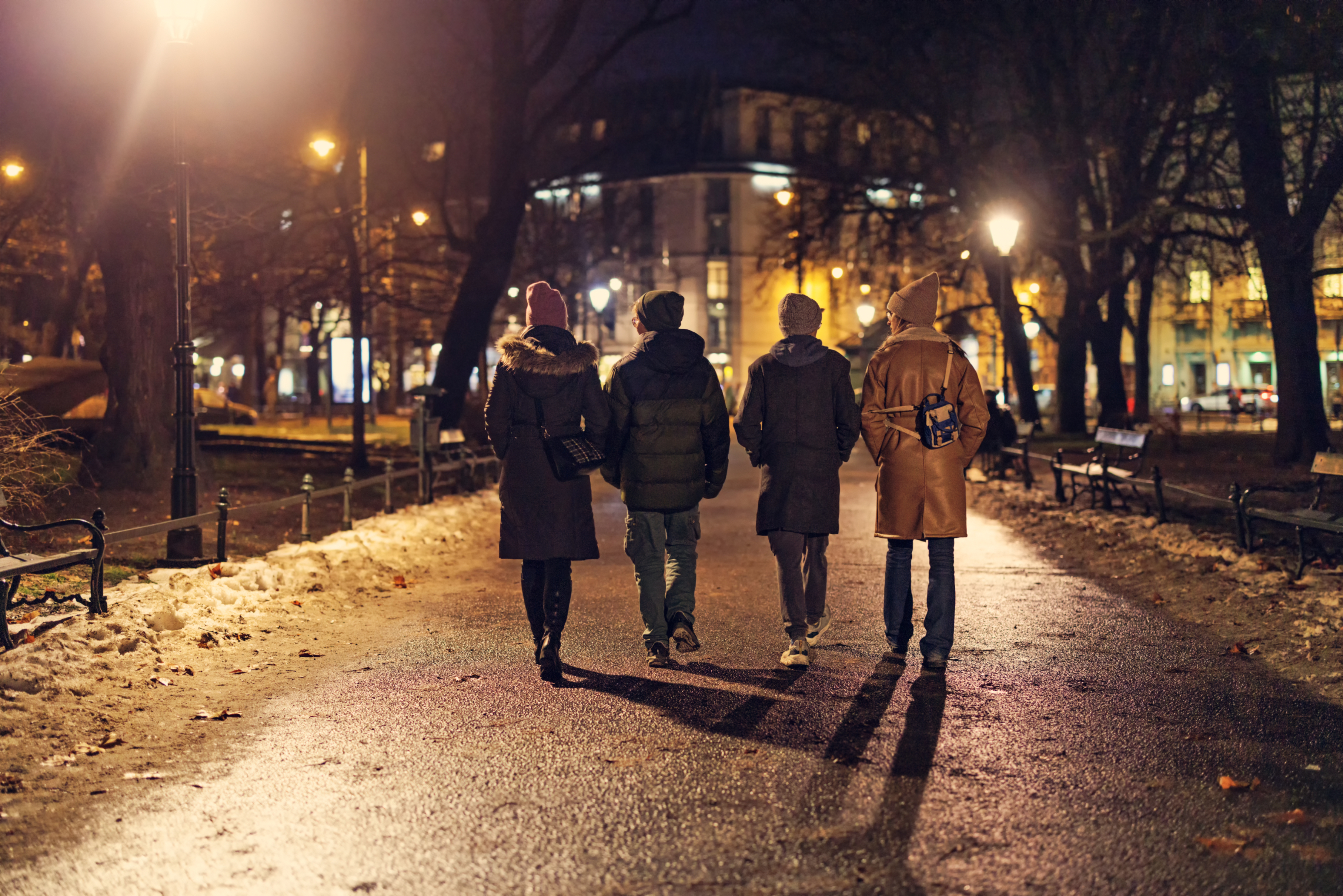 Four people are walking in a park at night, with their backs to the camera. They are dressed warmly in coats and hats