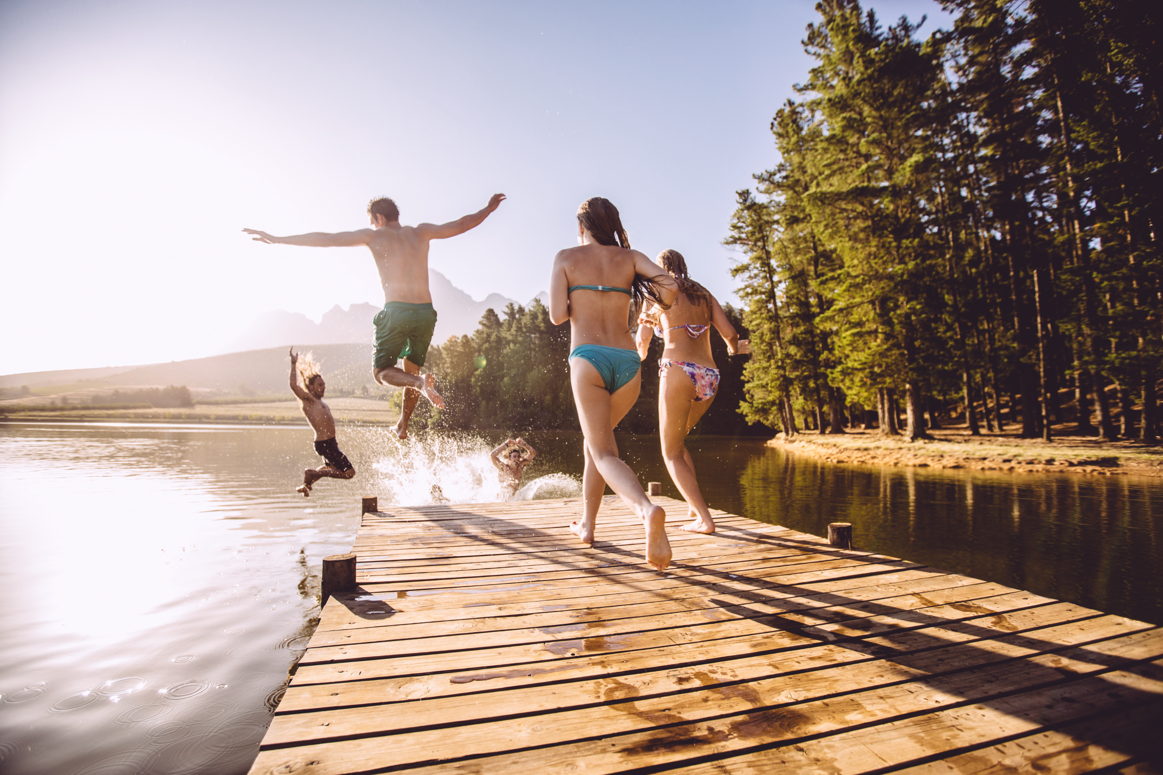 Four people jump off a wooden dock into a lake on a sunny day in a forested area