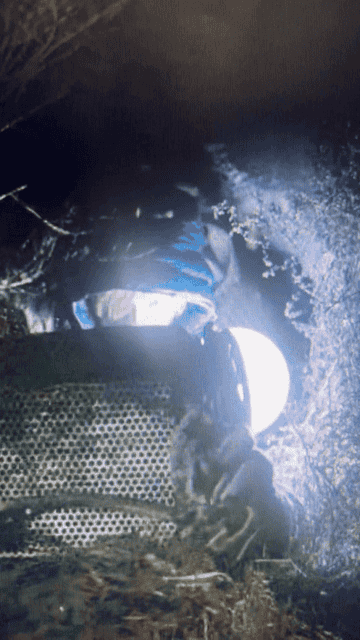 An astronaut in a spacesuit holds a helmet under a space capsule or spacecraft, with details partially obscured by shadows and ice