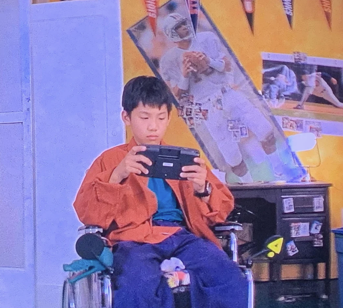 Boy sitting in a wheelchair playing a handheld game console in a sports-themed bedroom. Football posters are displayed on the wall behind him