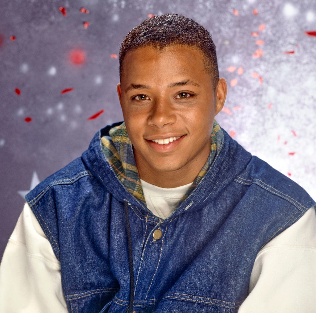 A young man, dressed in a casual denim jacket layered over a plaid hoodie, smiles against a festive background with confetti and sparkles