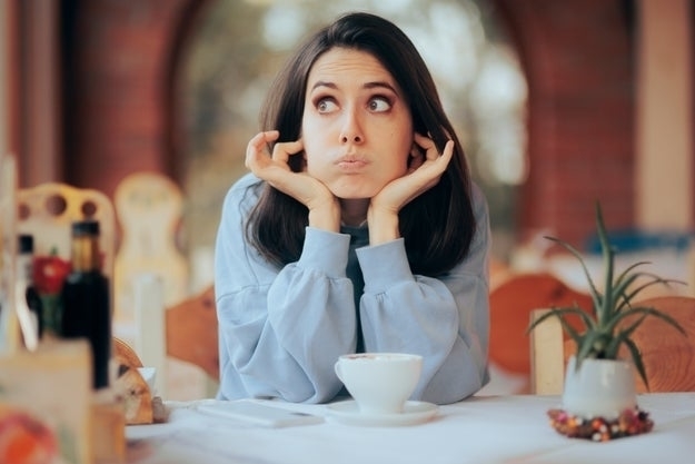 Woman with brown hair in a cafe, wearing a casual long-sleeve top, looks thoughtful with hands on her cheeks. A cup of coffee and a small potted plant are on the table