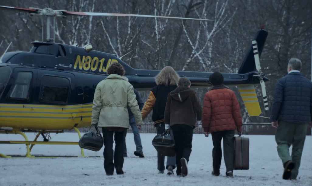 Five unidentified people walk towards a helicopter on a snowy ground, carrying bags and dressed in winter jackets