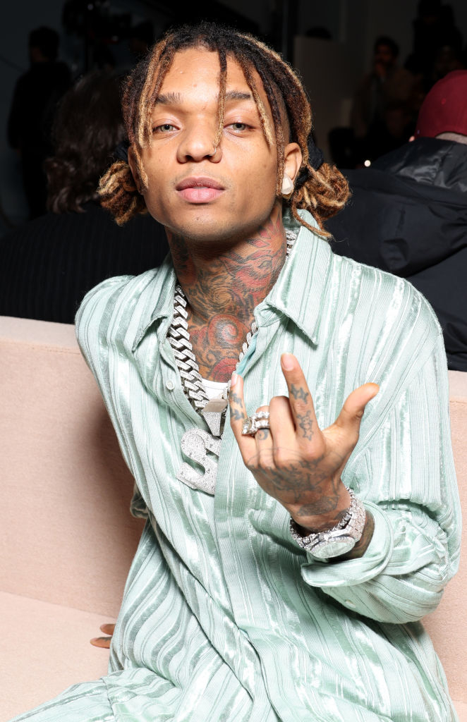 Swae Lee is sitting on a beige couch, wearing a light, patterned shirt and several necklaces, while holding up his left hand with rings on his fingers