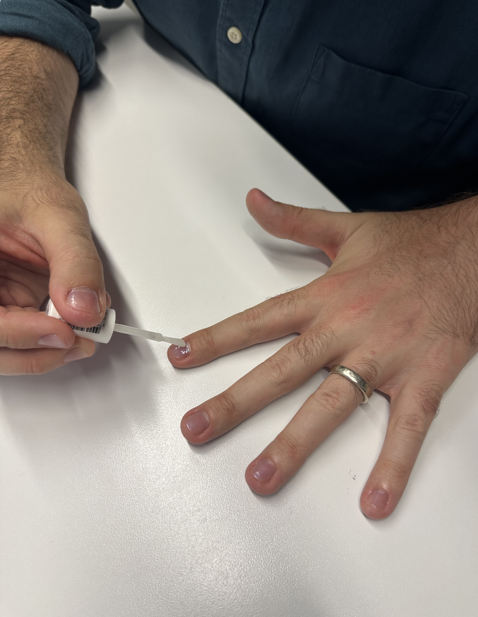 A person with a wedding ring applies clear nail polish to their fingernails on a white surface