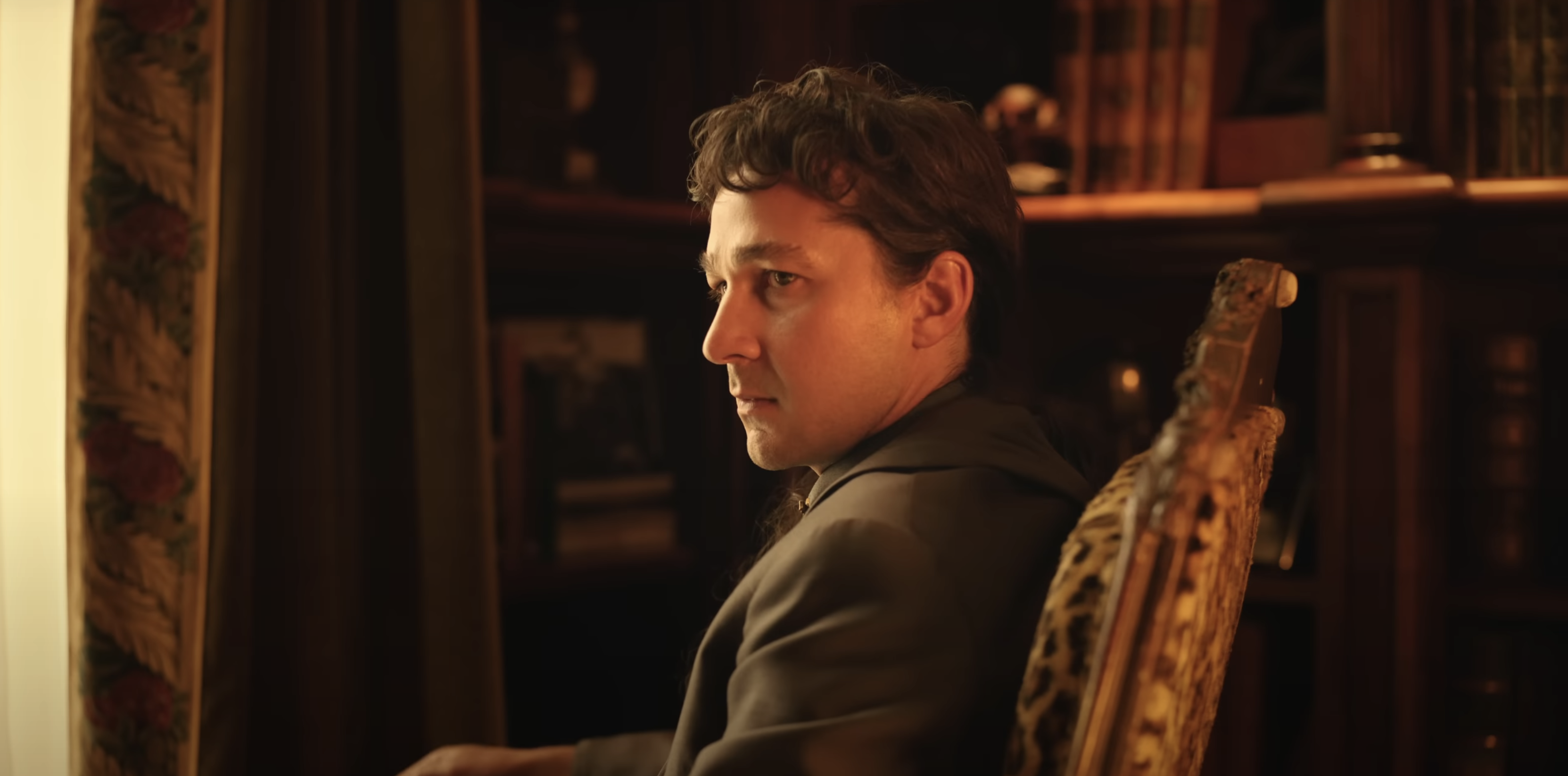 Shia LaBeouf in character, seated in an ornate chair, looking intently to the side within a dimly lit, vintage-styled room, likely from a movie scene