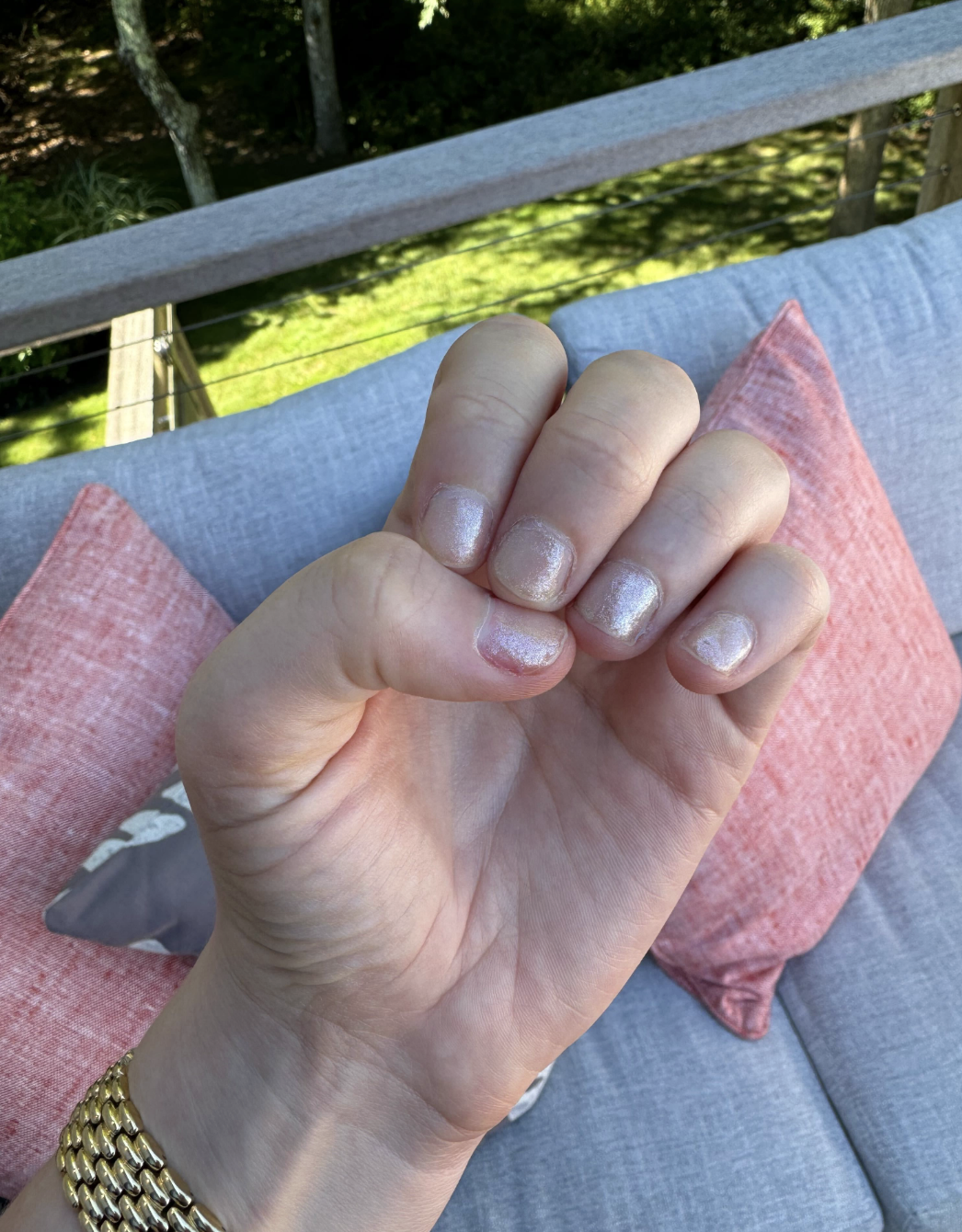 Close-up of a person's hand with neatly polished fingernails, wearing a gold bracelet, resting on a gray and pink cushioned outdoor chair